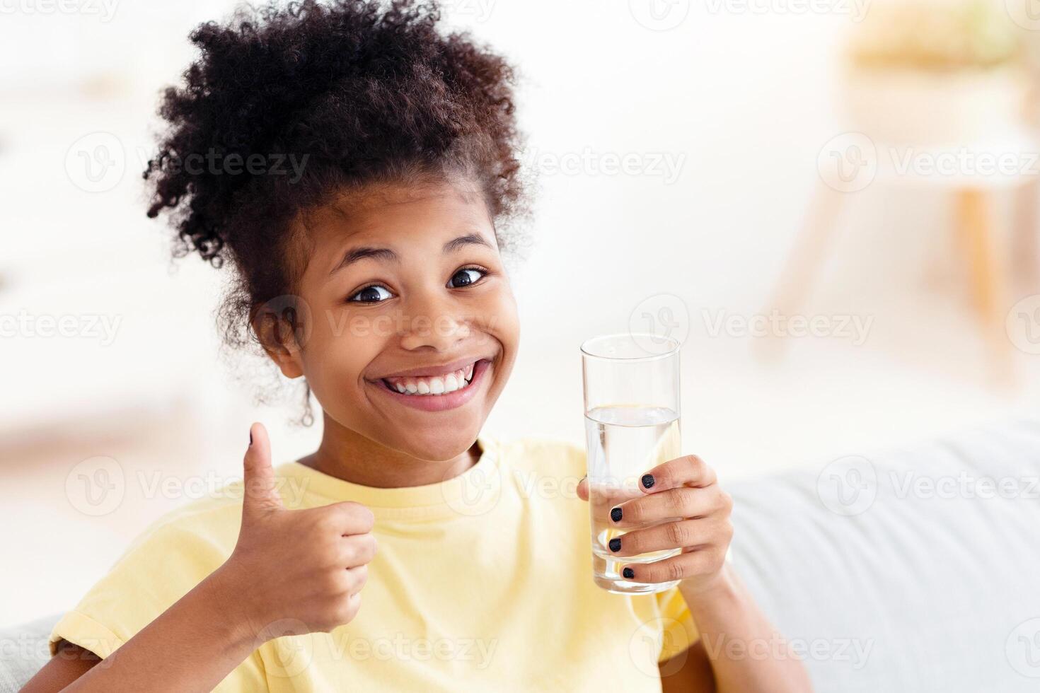 Hydration. Happy African Teen Girl Holding Glass Of Water Gesturing Thumbs Up Sitting On Couch At Home, Smiling To Camera photo