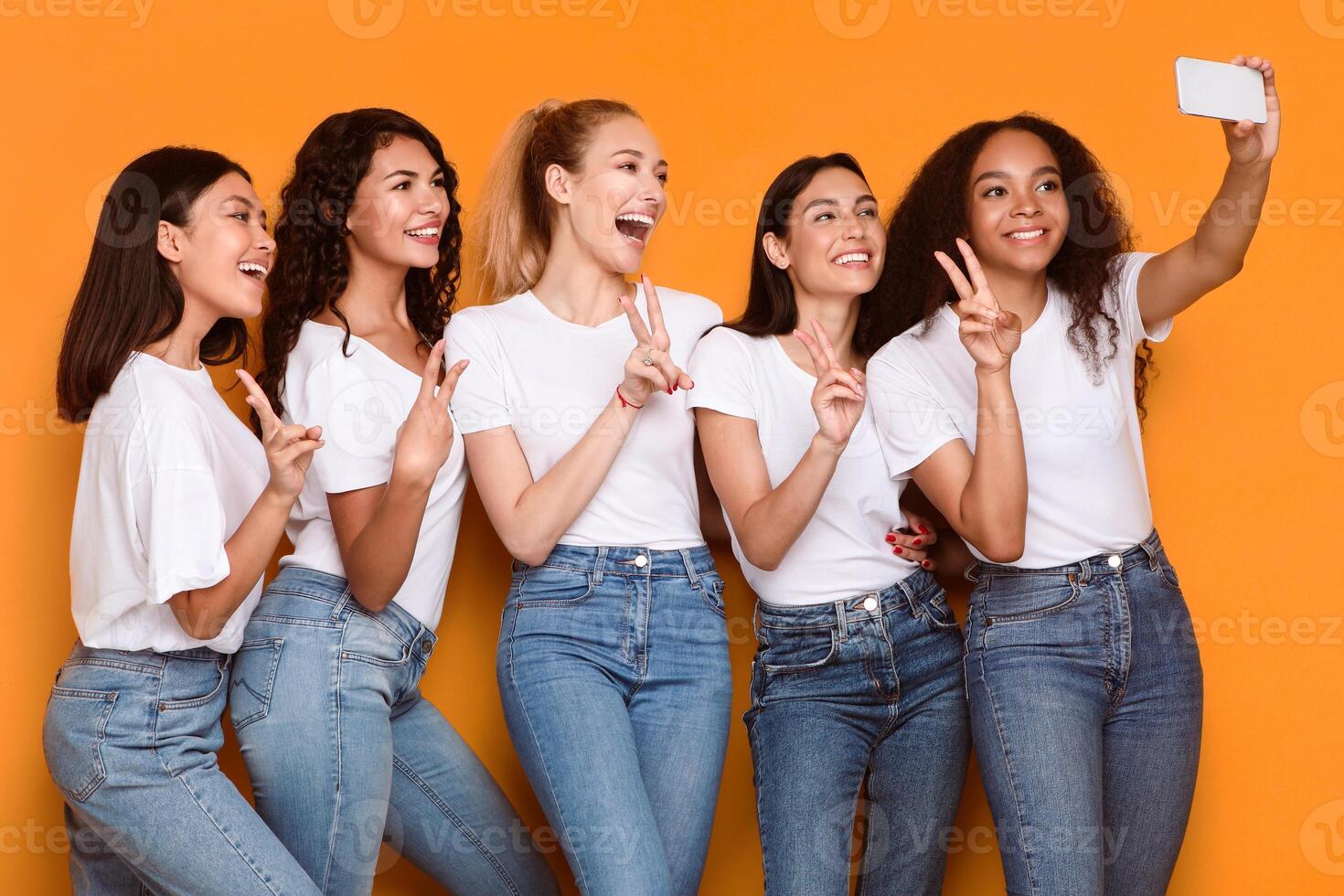 Multicultural Ladies Making Selfie On Phone Gesturing V-Sign Standing On Yellow Studio Background. Diverse Women Having Fun Posing And Taking Pictures On Smartphone. Female Friendship Concept photo