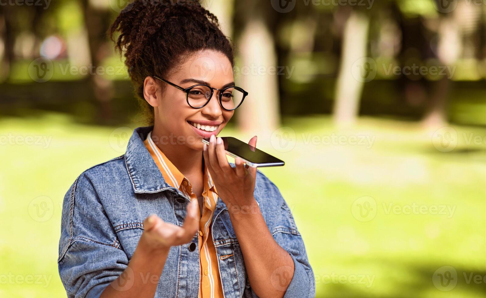 Happy Black Lady Talking On Cellphone Using Loudspeaker Mode Walking Outdoors In City Park. Smartphone User Chatting On Phone, Modern Mobile Communication Concept. Empty Space For Text photo