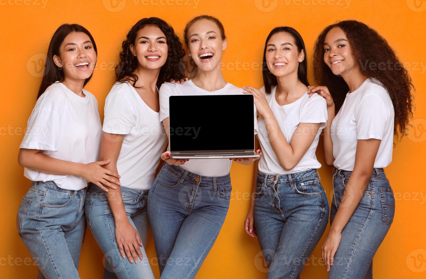 Five multiethnic women holding laptop showing blank computer screen recommending girlish website standing over yellow studio background. Mockup photo