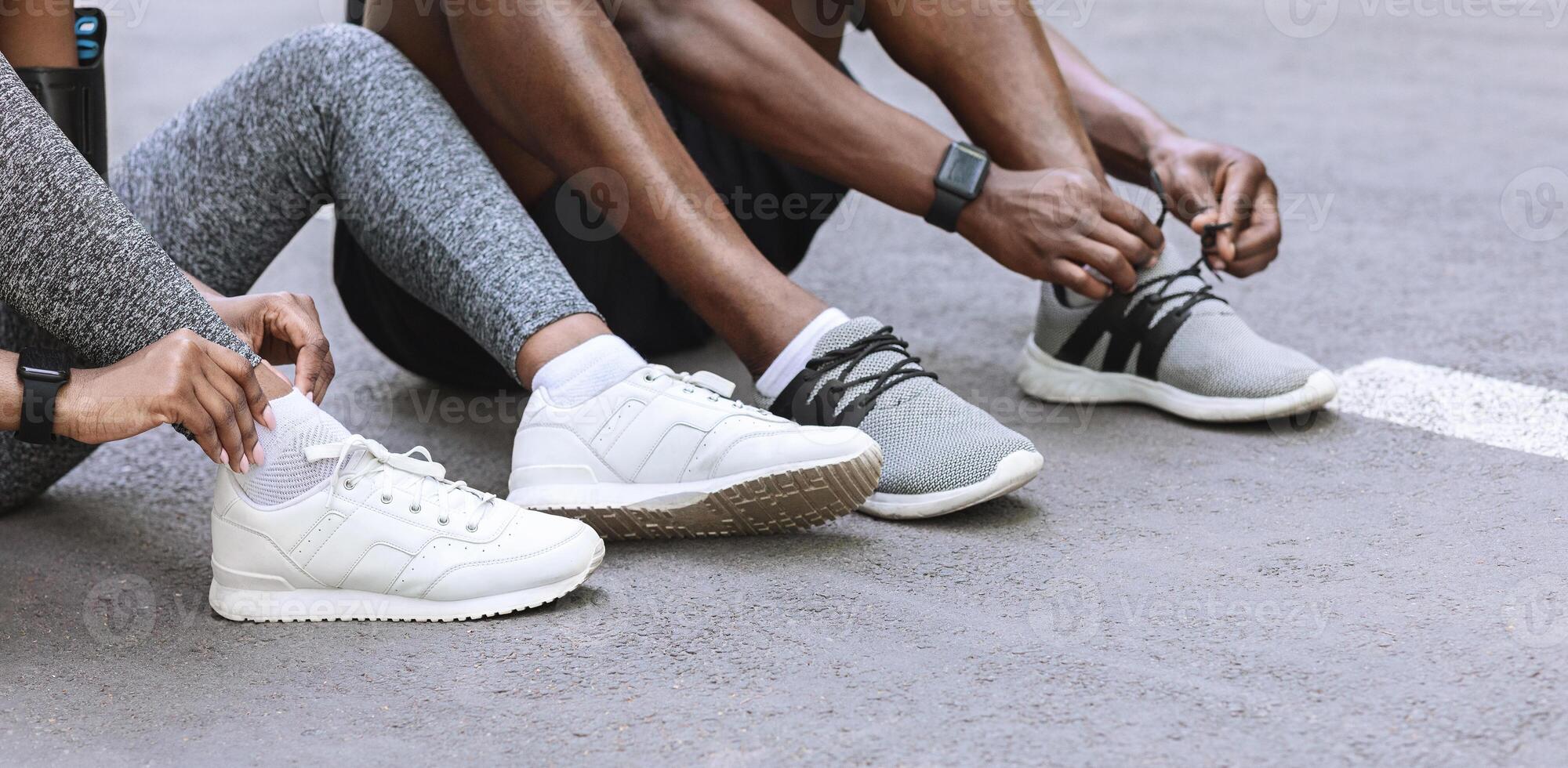 Couple of black runners lace their shoes outdoors, preparing for jogging, sitting on asphalt path, cropped image with copy space photo