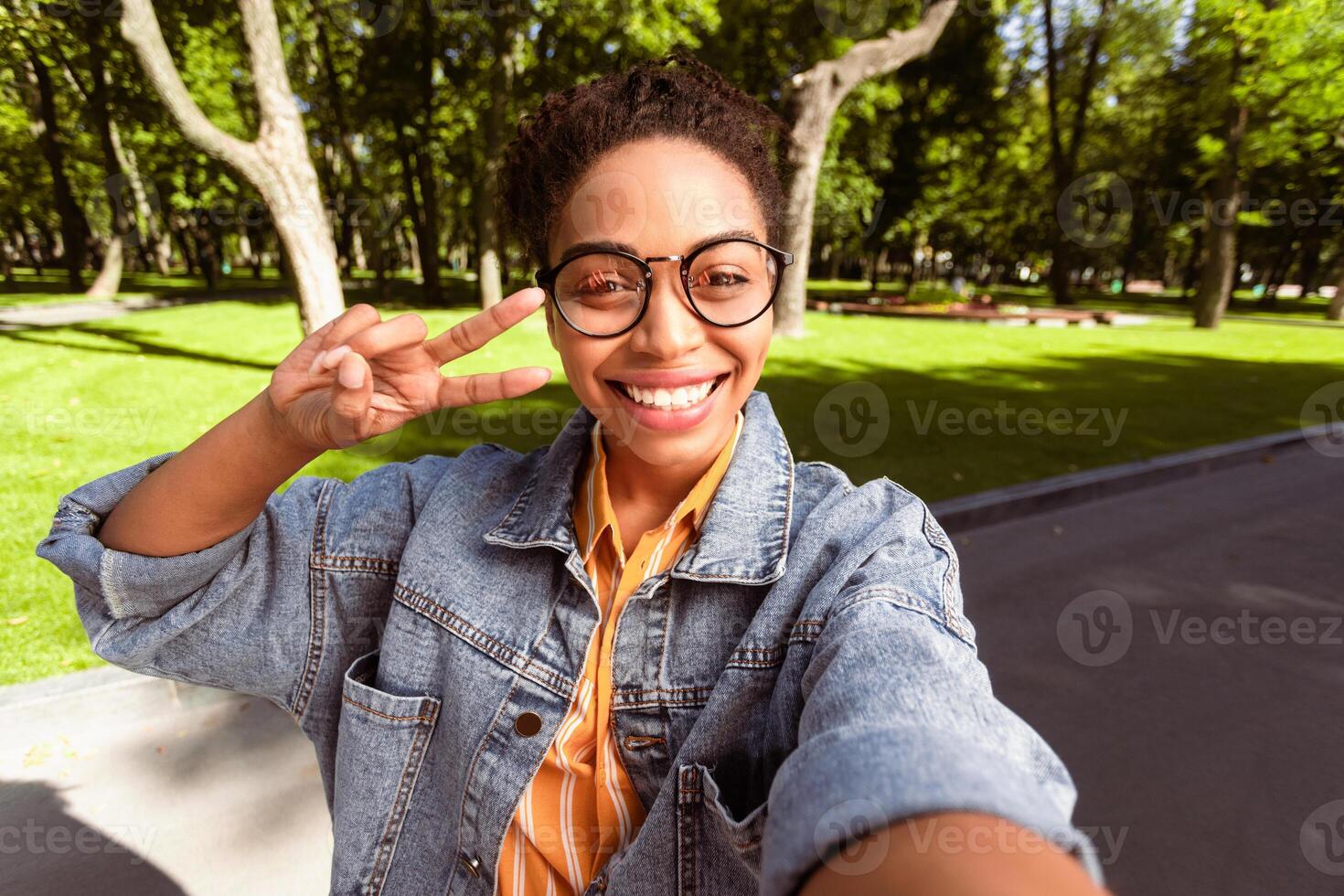 Cheerful Black Female Student Making Selfie Gesturing V-Sign Having Fun, Smiling To Camera Posing Outside Standing In Park. Self-Portrait, Mobile Photo Concept. Selective Focus