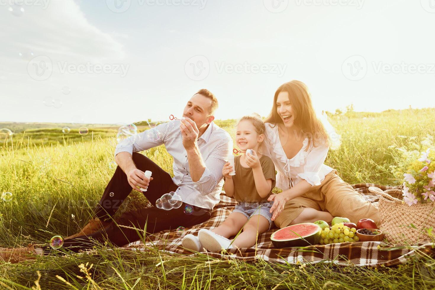 Outdoor Activity Concept. Young parents blowing soap bubbles with their child, having family picnic, copy space photo