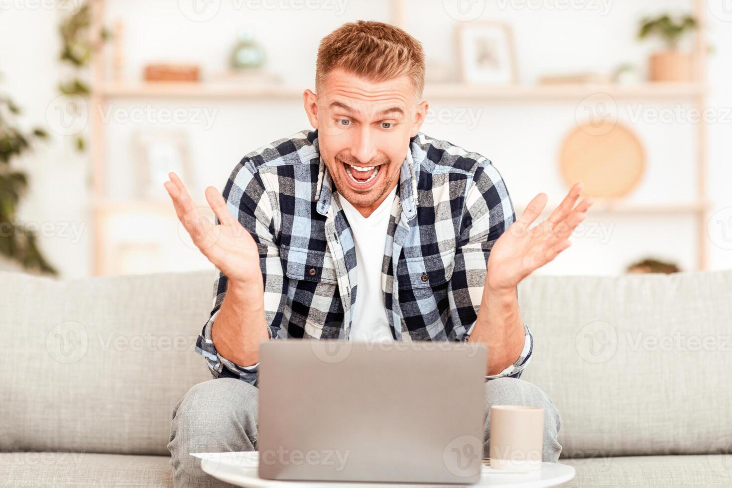 Online Win Concept. Portrait of happy excited man celebrating his success sitting on the couch in living room, screaming with joy, looking at laptop, free space. Amazed guy using computer photo