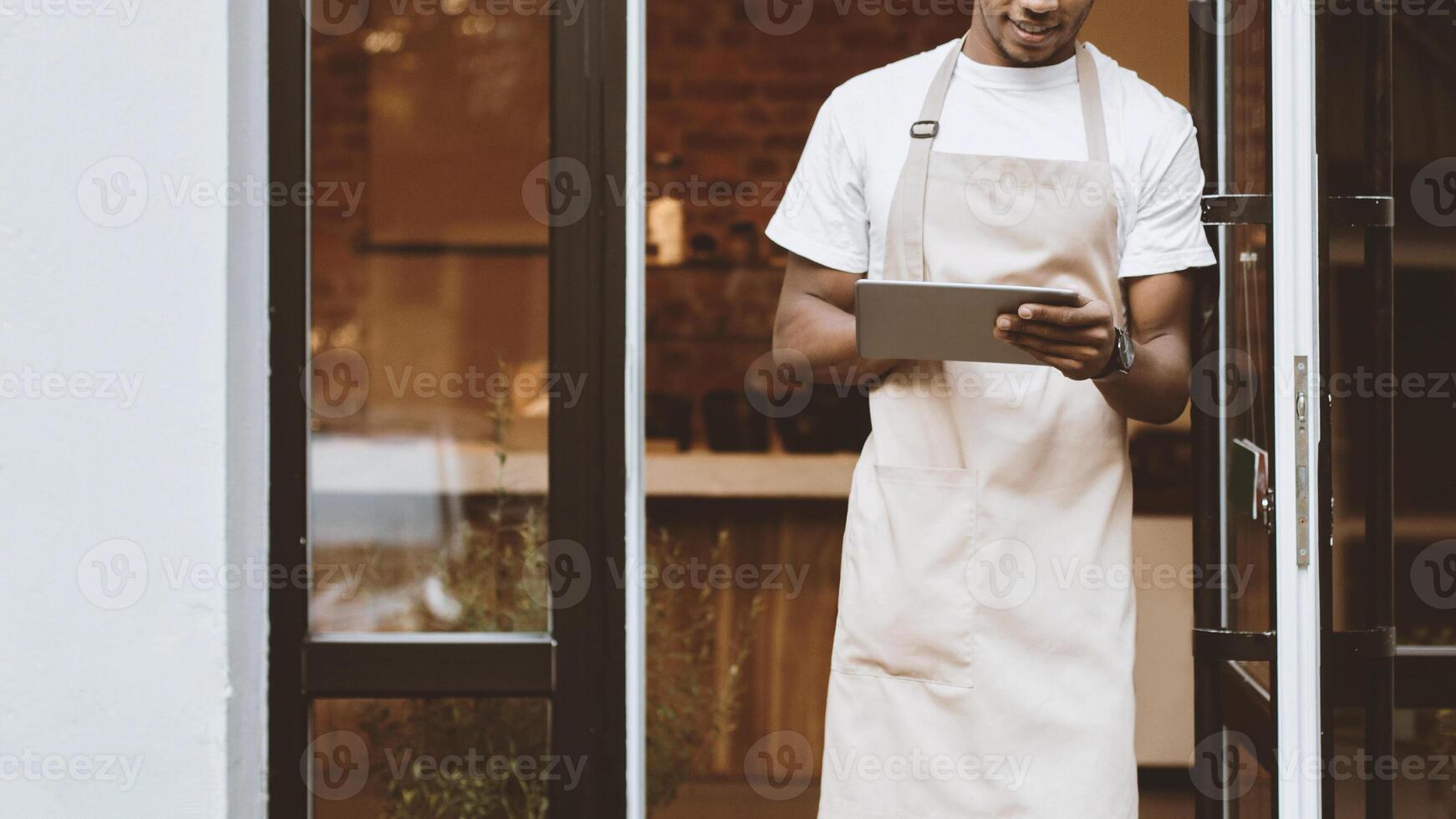 New normal and ordering coffee online for takeaway during covid-19 quarantine. Millennial african american male in apron works on tablet near front door of modern cafe, cropped, empty space photo
