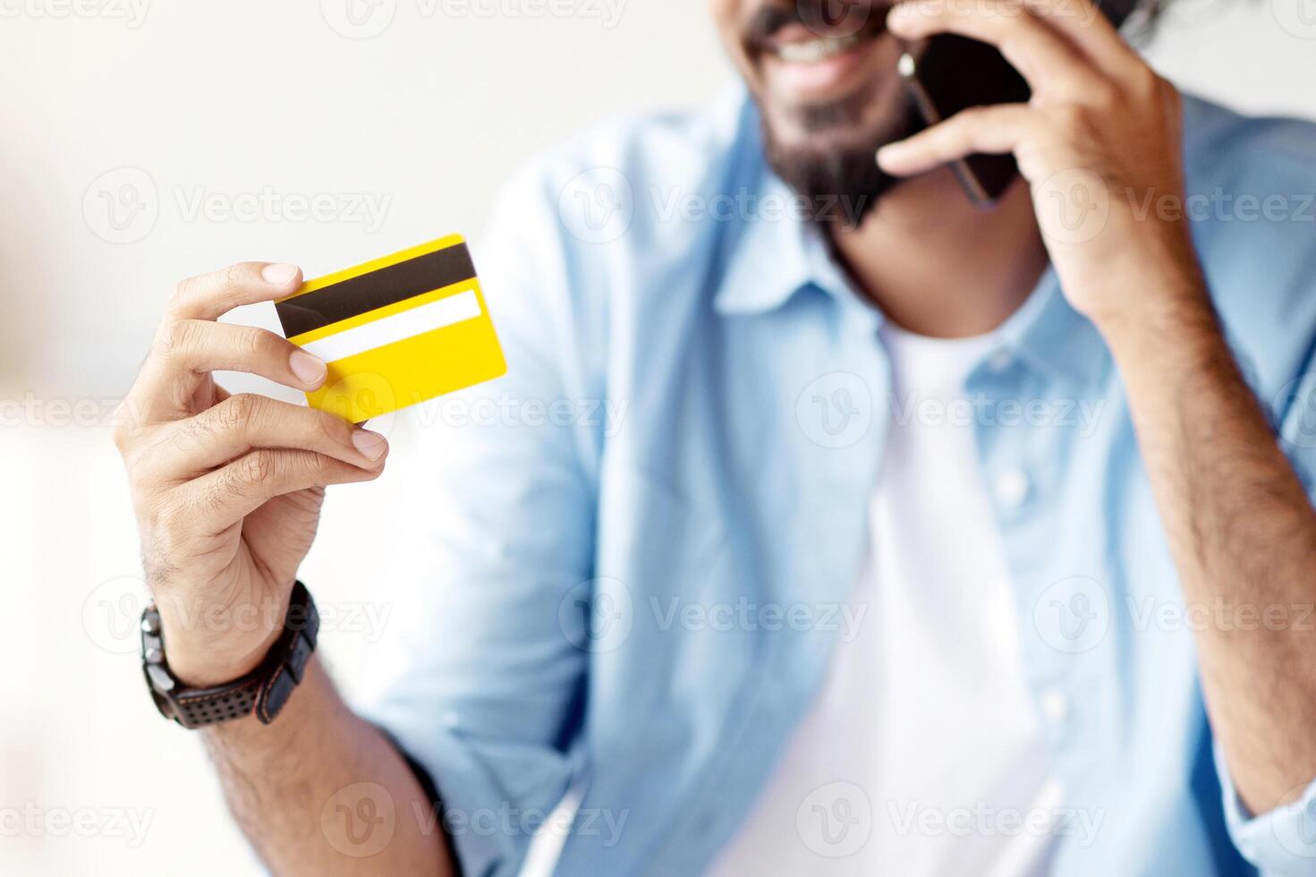 Mobile Payment. Positive Indian Man Holding Credit Card And Talking On Cellphone In Office, Transfering Money, Using Online Banking, Talking With Customer Support, Paying In Internet, Selective Focus photo