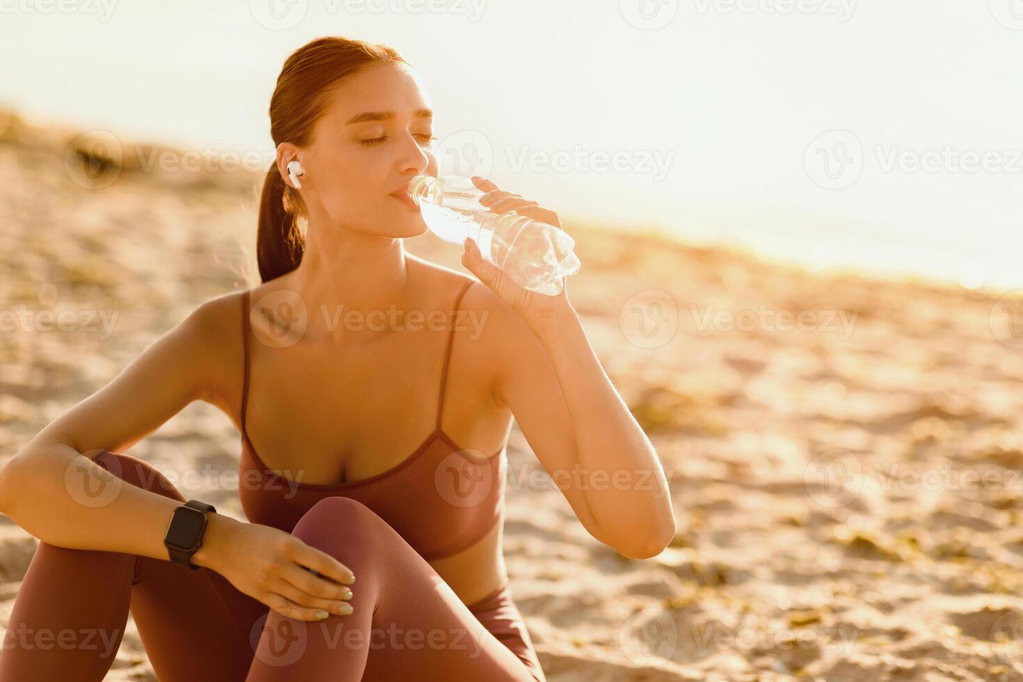 Break From Work Out. Slim woman in sports bra and earbuds drinking water, resting on the seaside, copy space photo