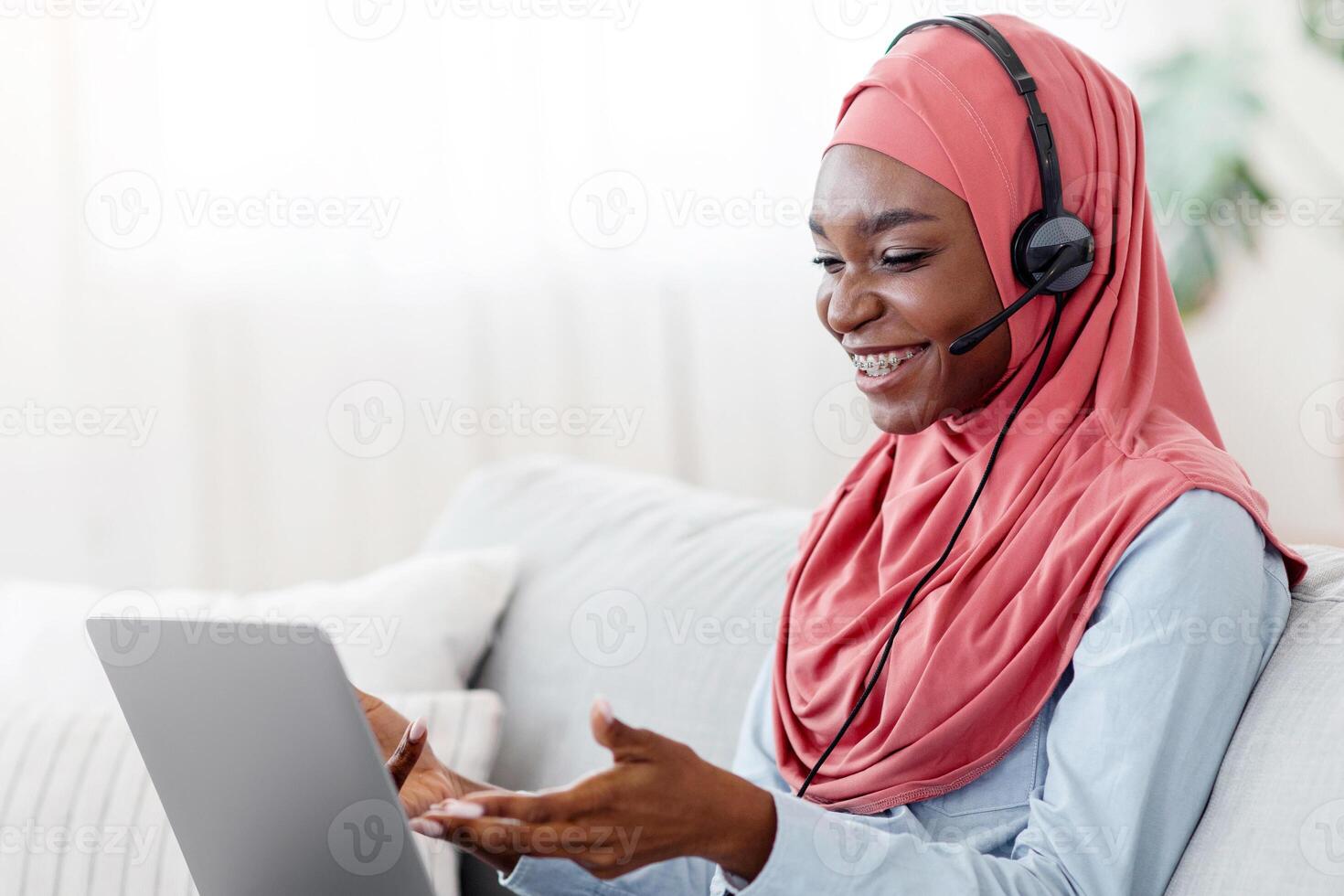 Online business meeting. Smiling black muslim lady in hijab using headset and laptop at home, having conference, sitting with computer on comfortable couch in living room, talking on pc camera photo