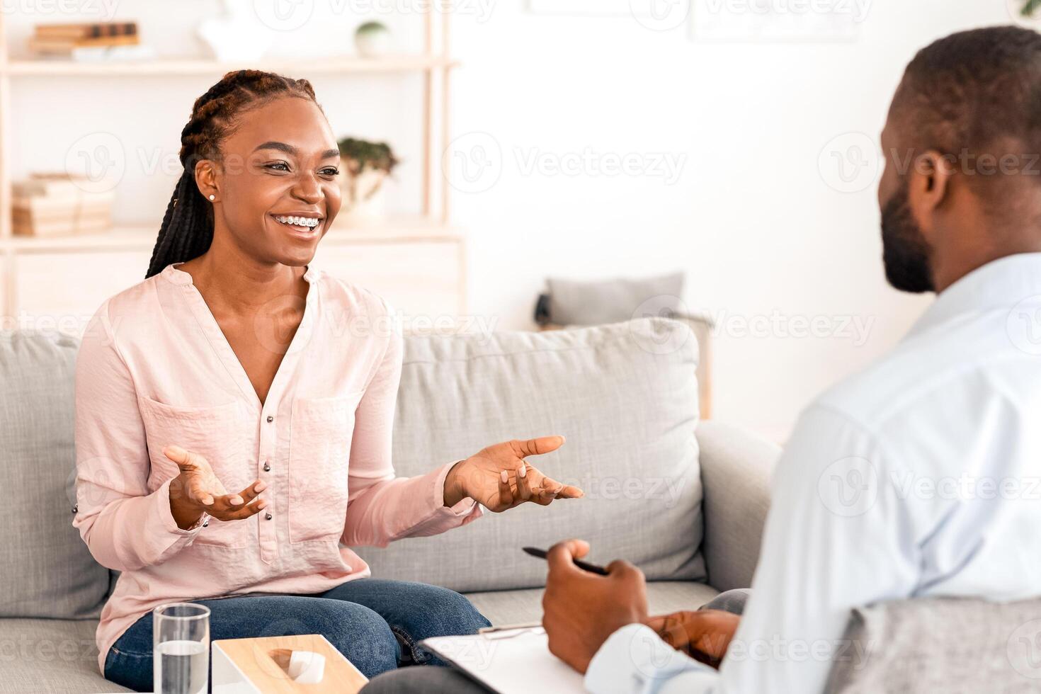 Happy african woman talking with psychologist at private therapy session, sharing her progress, sitting on couch and smiling, free space photo