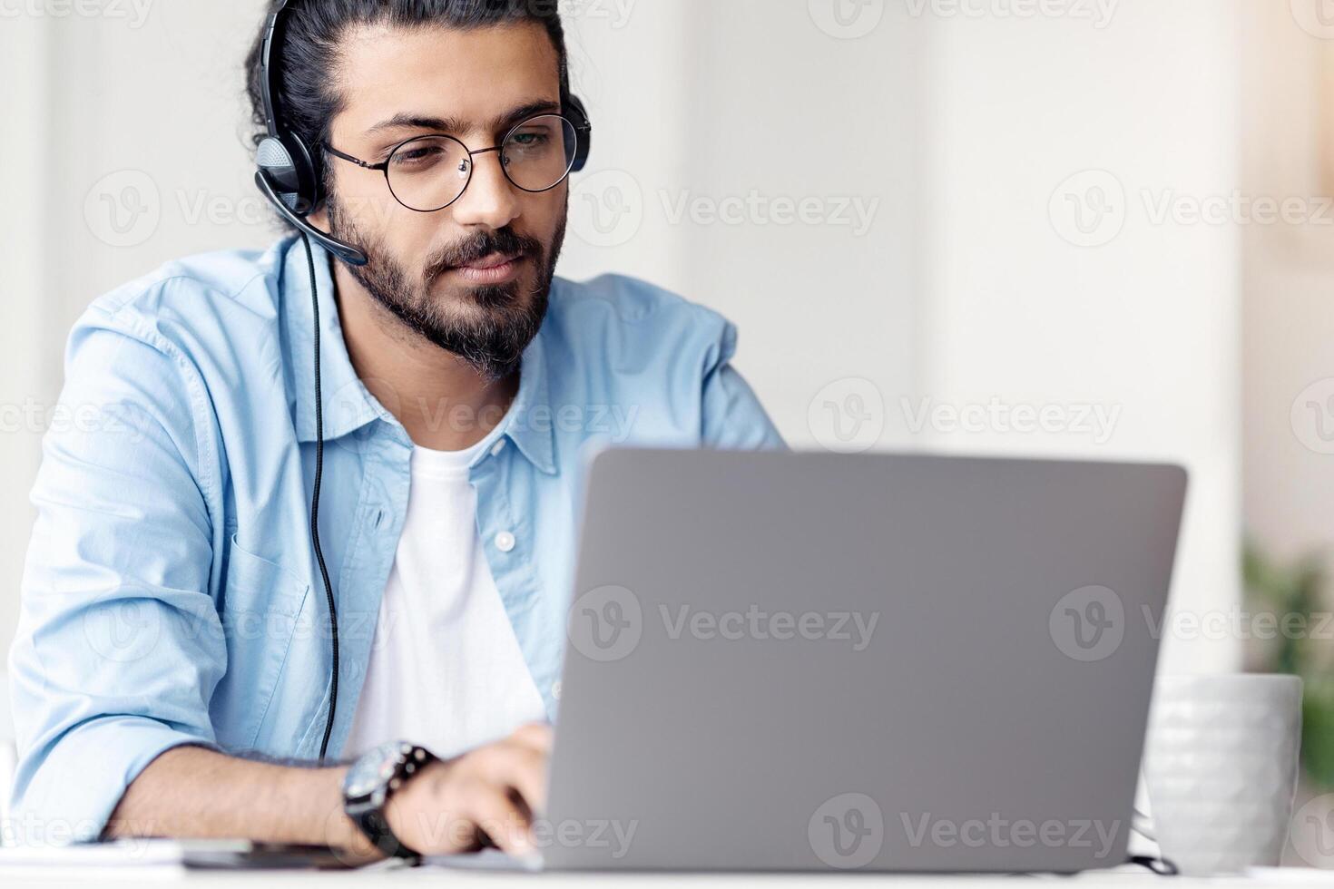 Young Arab Male Hotline Operator In Headset Working With Laptop In Modern Office, Handsome Eastern Guy Customer Support Manager Sitting At Desk And Typing On Computer, Closeup Shot photo