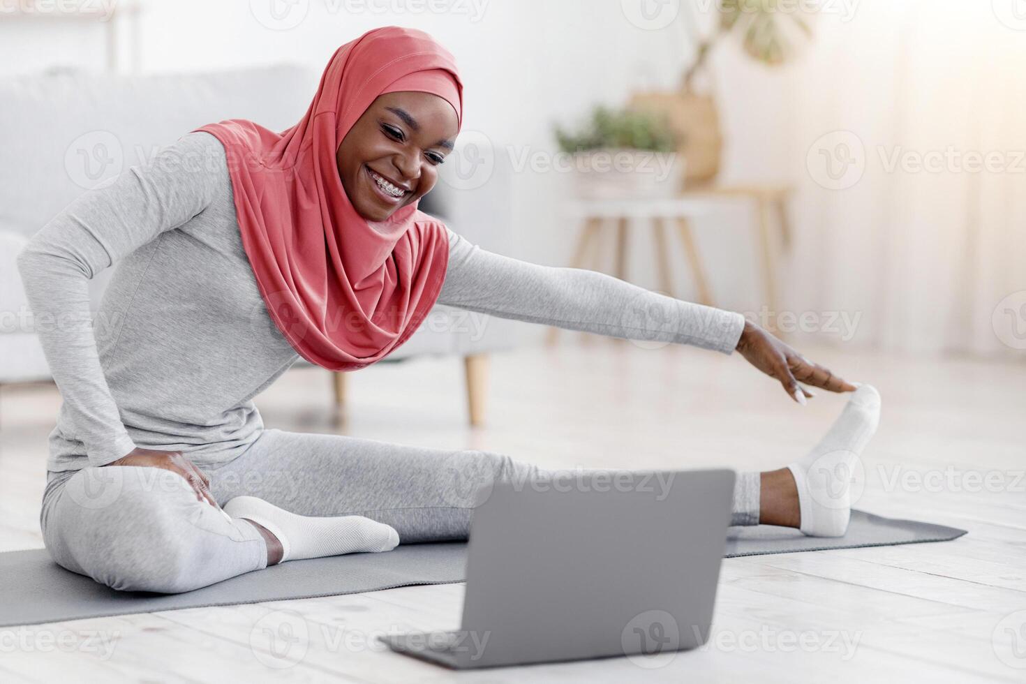 Sporty young black muslim woman in hijab doing stretching exercises in front of laptop at home, training in living room, watching online tutorials, religious african woman enjoying healthy lifestyle, photo