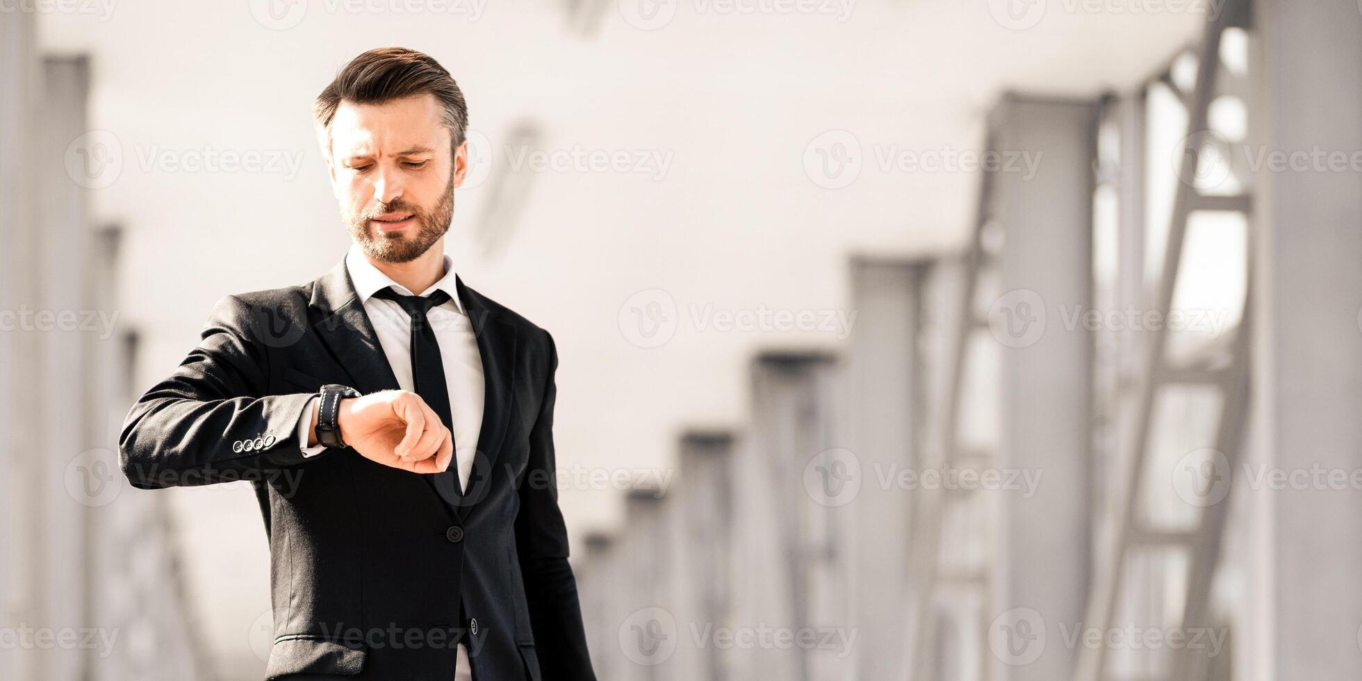 Worried middle-aged businessman walking by airport terminal, carrying luggage and checking time, looking at watch, panorama with copy space. Stressed entrepreneur in suit getting late for flight photo