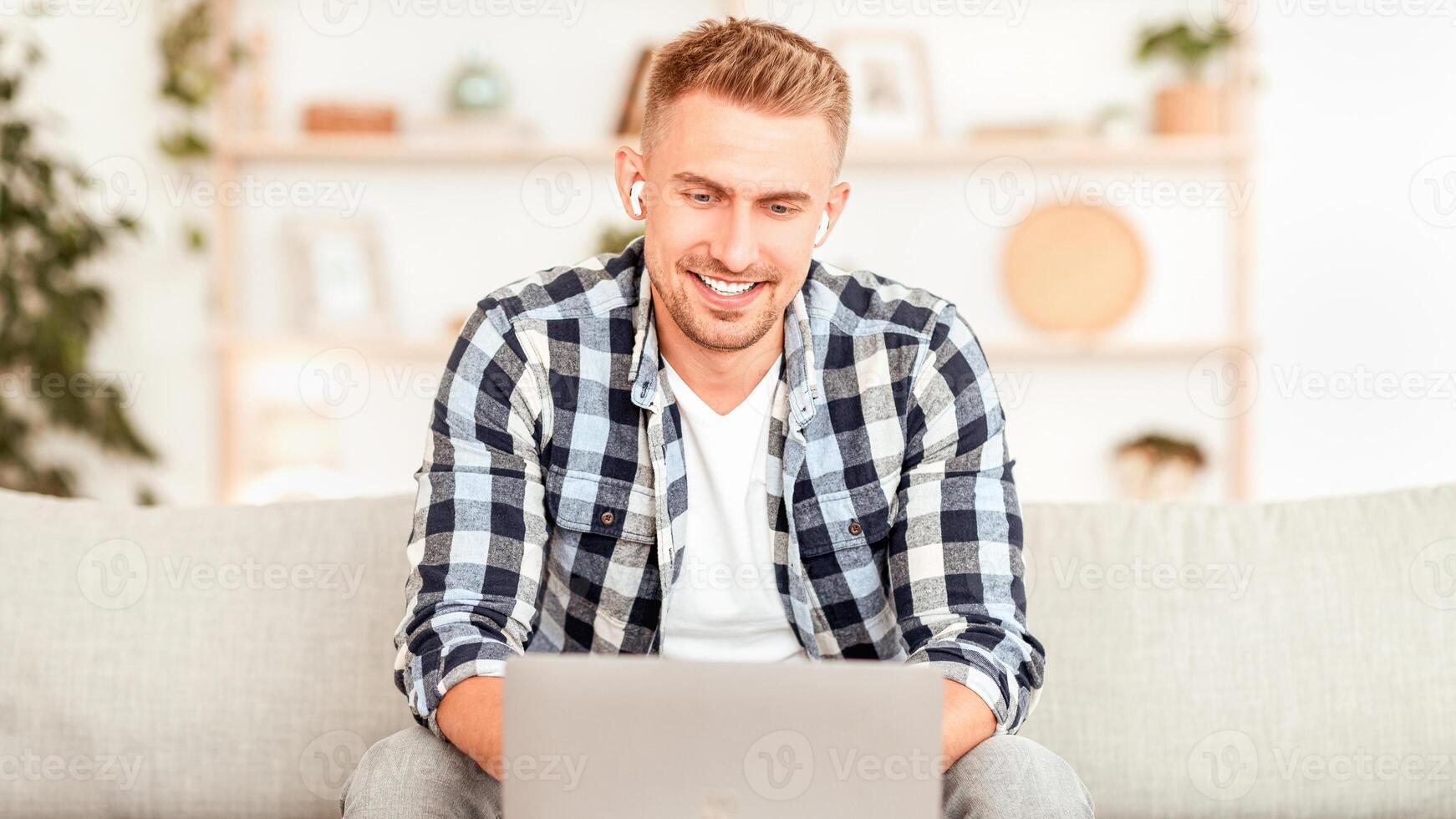 Lifestyle Concept. Portrait of smiling man working online, using computer sitting on the couch and wearing wireless earphones, free space. Guy studying with music, listening to podcast or audio book photo