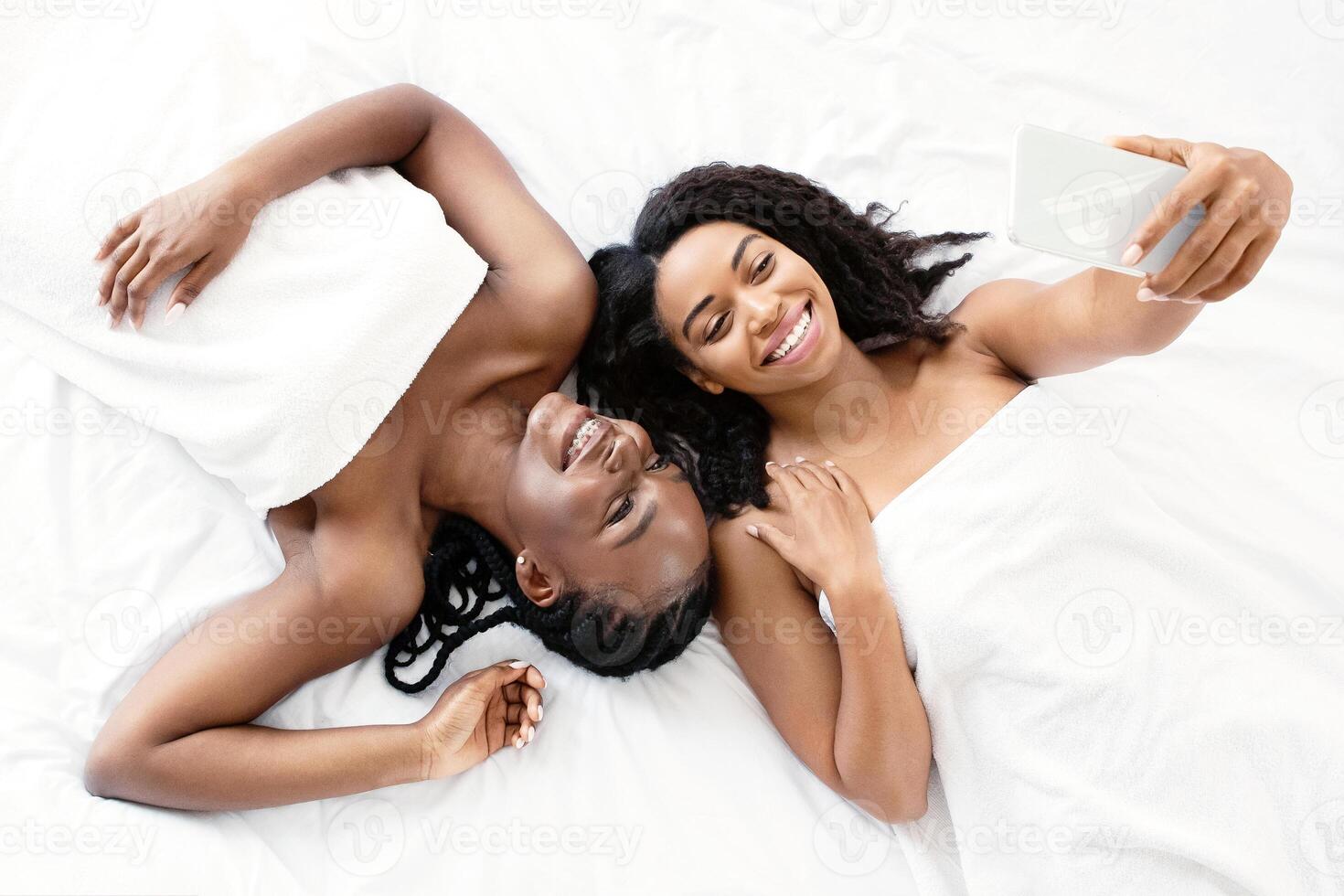 Beautiful topless african american girlfriends taking selfie on mobile phone, laying on bed covered in blankets, top view. Smiling black ladies laying head to head on bed, taking photo on smartphone