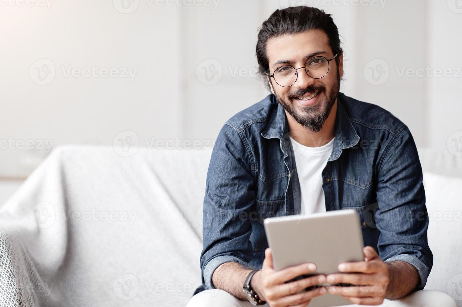 Handsome Indian Guy Holding Digital Tablet Computer And Smiling At Camera, Sitting On Couch At Home, Using Modern Technologies For Leisure, Browsing Internet Or Checking Social Networks, Free Space photo