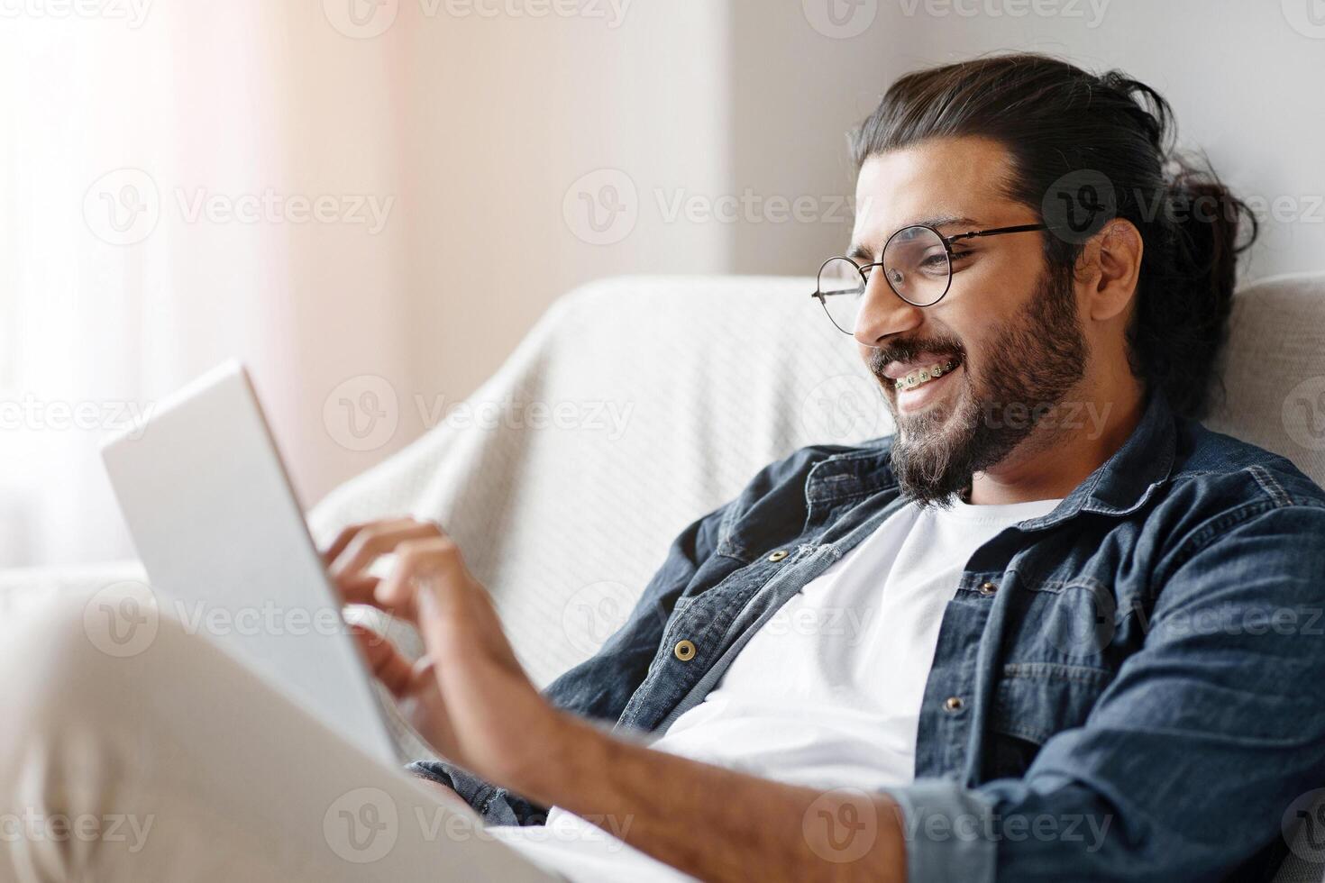 Happy western man checking social media on digital tablet at home, sitting on comfortable couch in living room, browsing internet or new app, enjoying domestic passtime and modern technologies photo