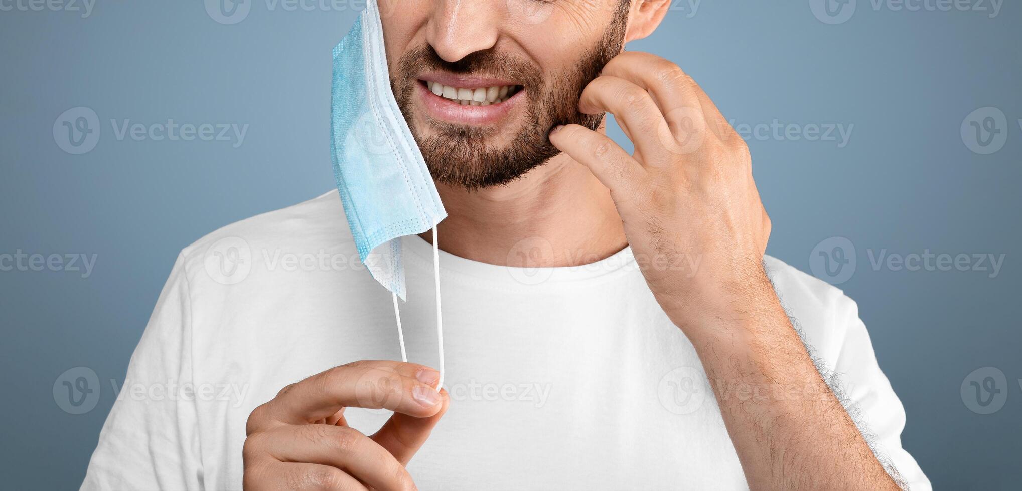Bearded man moved protective medical mask from face to scratch beard, grey studio background, free space. Do not touch your face, avoid touching your face during pandemic, stay safe during lockdown photo