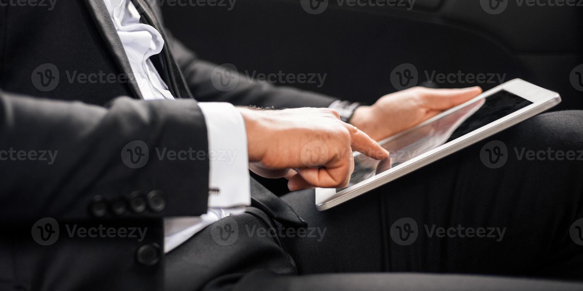 Modern technologies in business. Cropped of businessman in suit sitting in car, using digital tablet, working online. Unrecognizable man working on digital pad while going to airport, side view photo