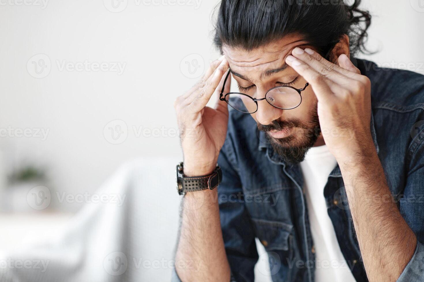 Young indian guy freelancer suffering from headache after hard working day, sitting on couch at home, massaging head temples, having strong migraine, feeling tired, upset and stressed, copy space photo