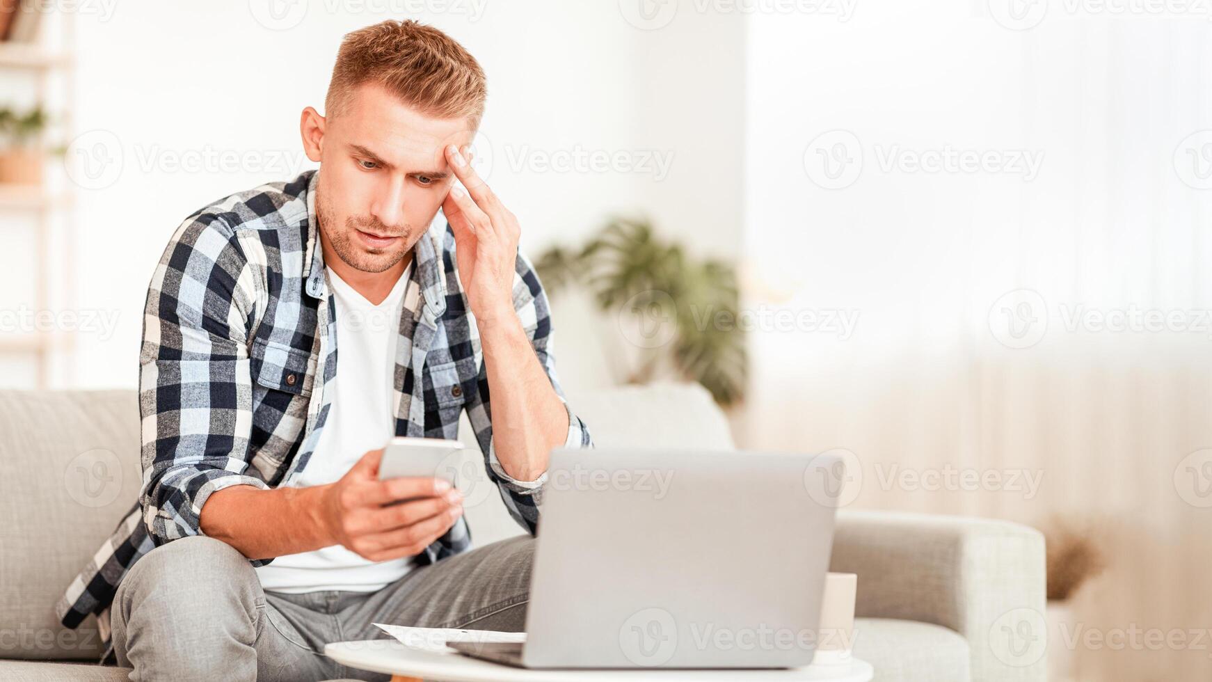 Unpleasant Message Concept. Portrait of upset depressed guy using his cellphone, disappointed and shocked by fake news, sitting on the sofa at home. Frustrated man looking at mobile phone photo