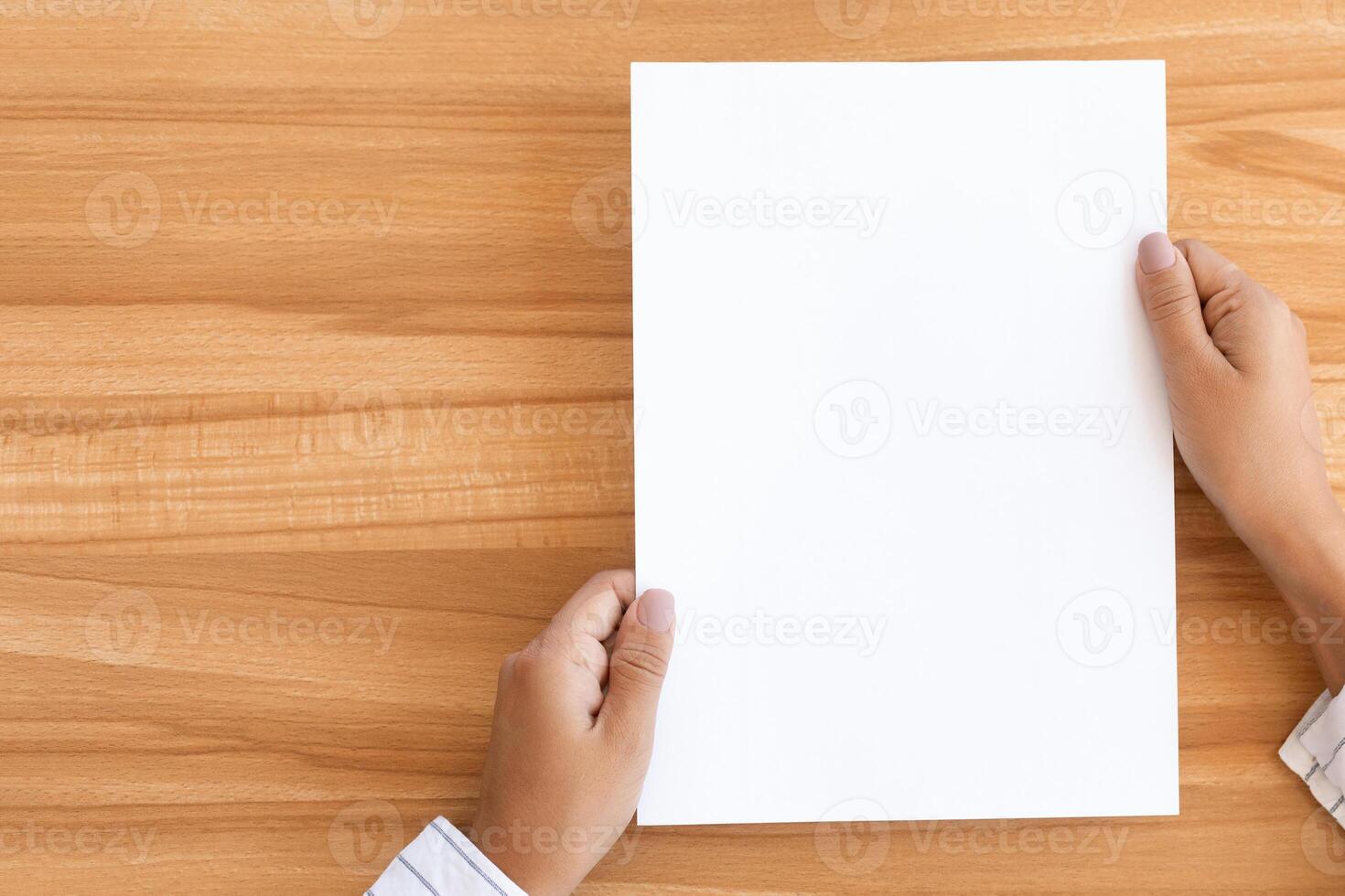 Overhead view of woman holding blank paper sheet A4 size or letter paper on wooden desk background, copy space photo