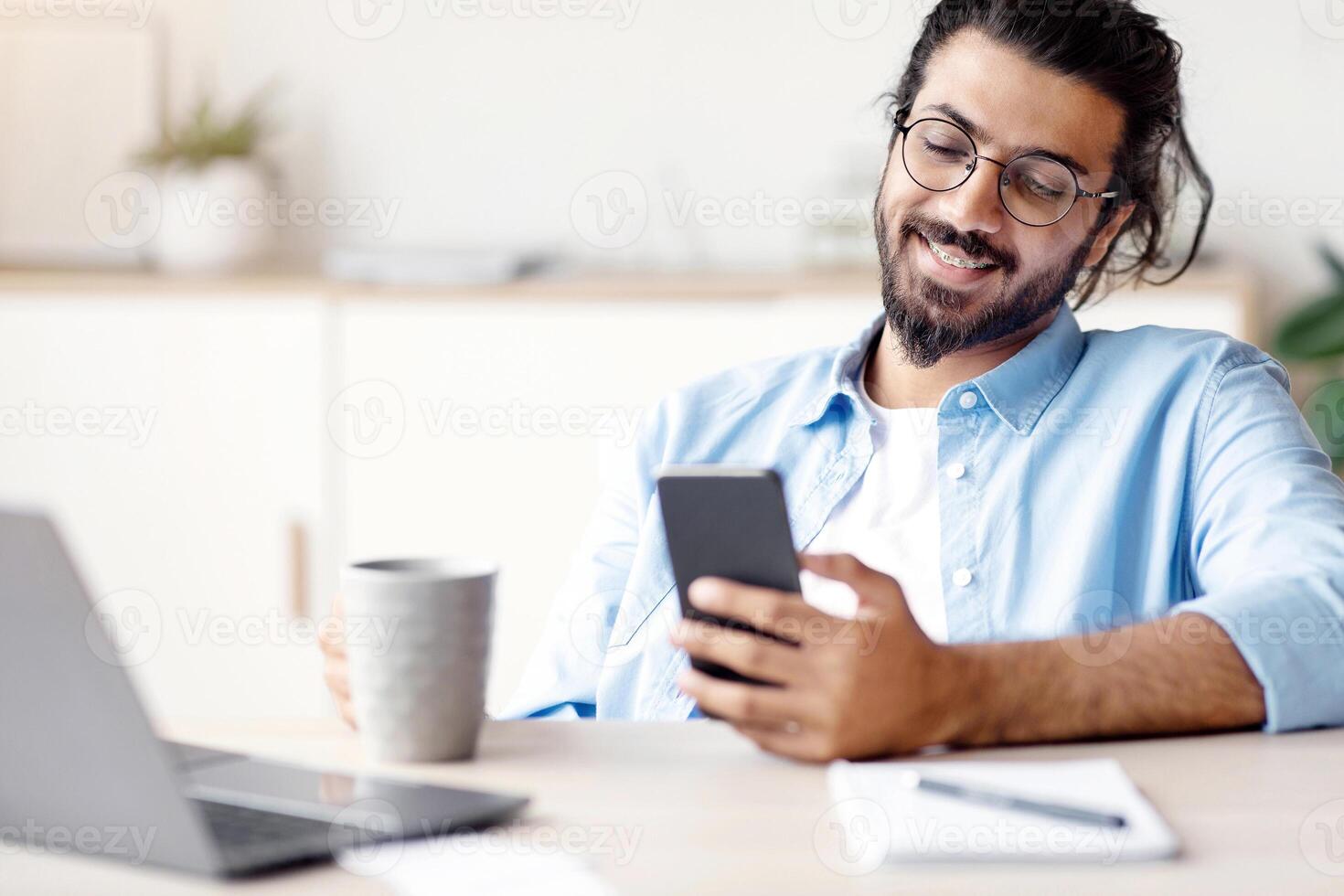Smiling Indian Employee Using Smartphone And Having Coffee In Office, Enjoying Break At Work, Texting With Friends Or Browsing Social Networks, Sitting At Desk With Laptop, Selective Focus, Free Space photo