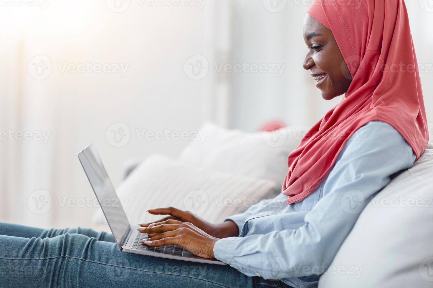 Muslim african woman in hijab using laptop at home, working remotely while sitting on comfortable sofa in living room, enjoying freelance online job, browsing internet, typing on keyboard, side view photo