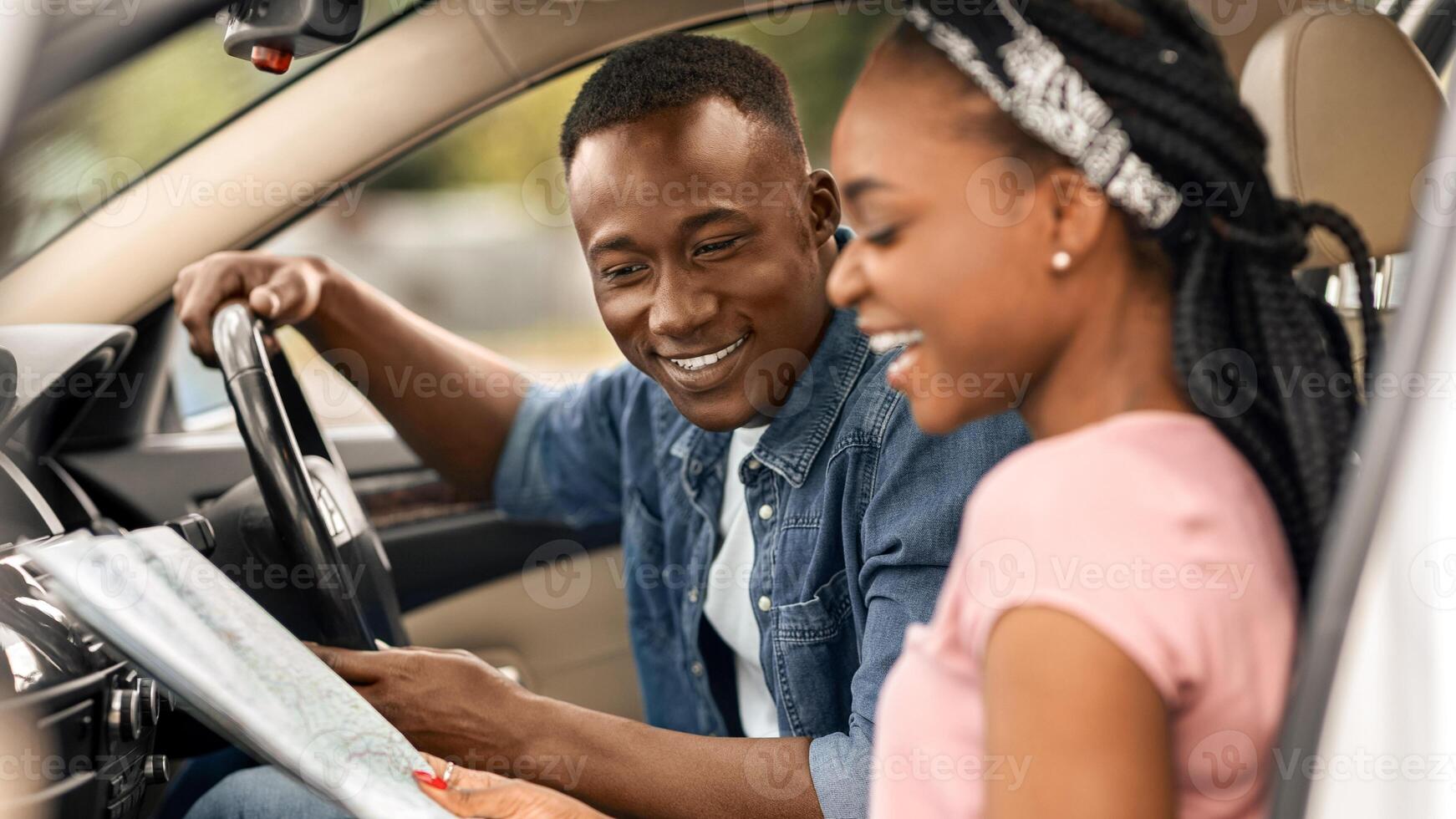 Car trip, travelling by auto concept. Happy millennial black woman showing her boyfriend destination to travel on paper map, sitting at front seats in automobile, discussing future journey, side view photo