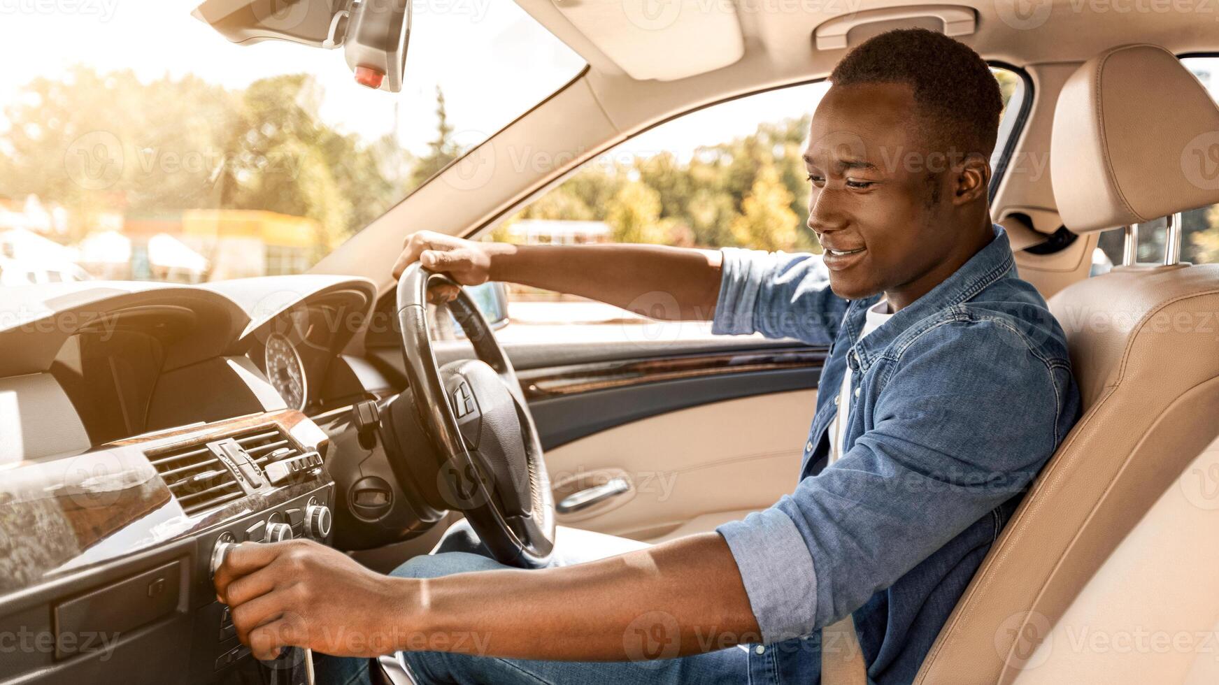 Excited young black man in casual sitting in comfy automobile, turning on music on dashboard before moving to office or beginning car trip, enjoying his new auto, side view, copy space photo