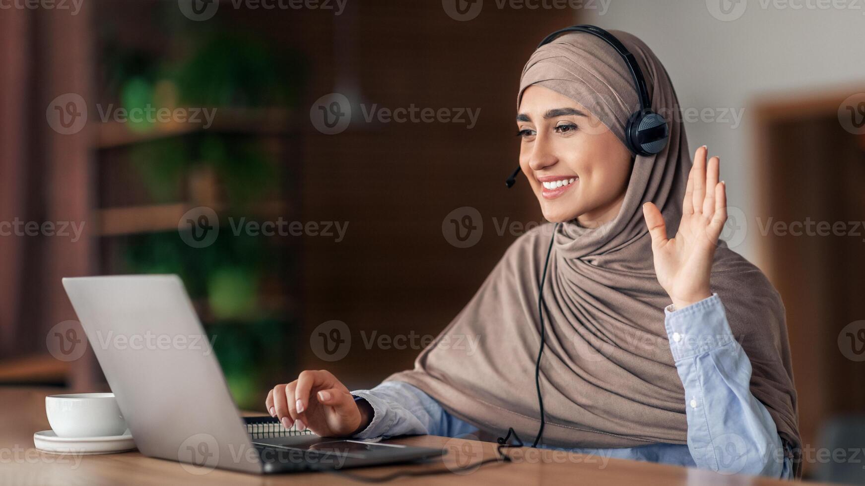 Smiling arab woman having call while resting at cafe, using laptop and headset, empty space. Cheerful young lady in hijab waving at laptop screen, attending conference, drinking coffee photo