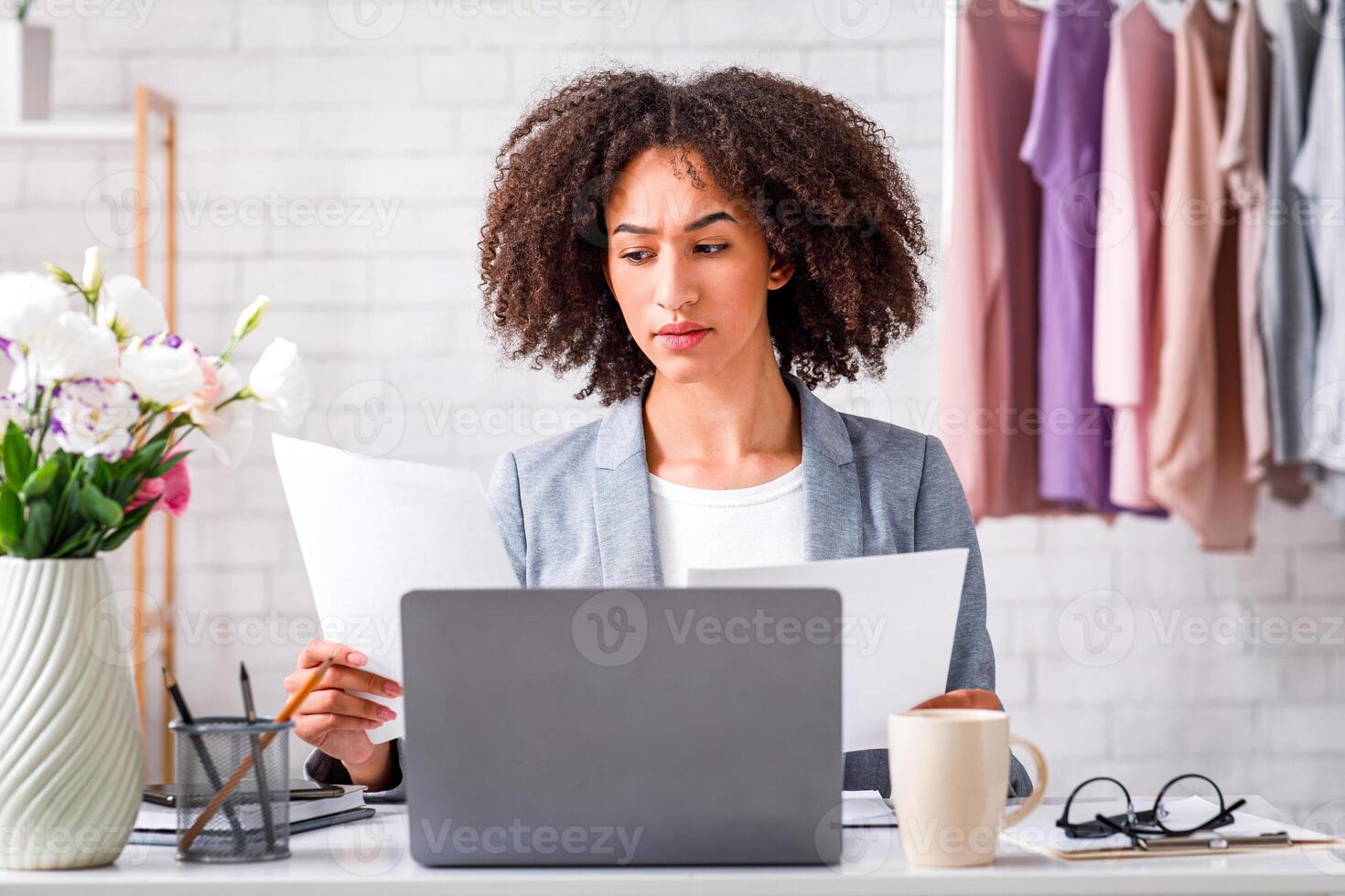 Paperwork and small business at home during coronavirus. Focused african american woman working with documents, sitting at table with laptop in interior with rack with clothes, free space photo