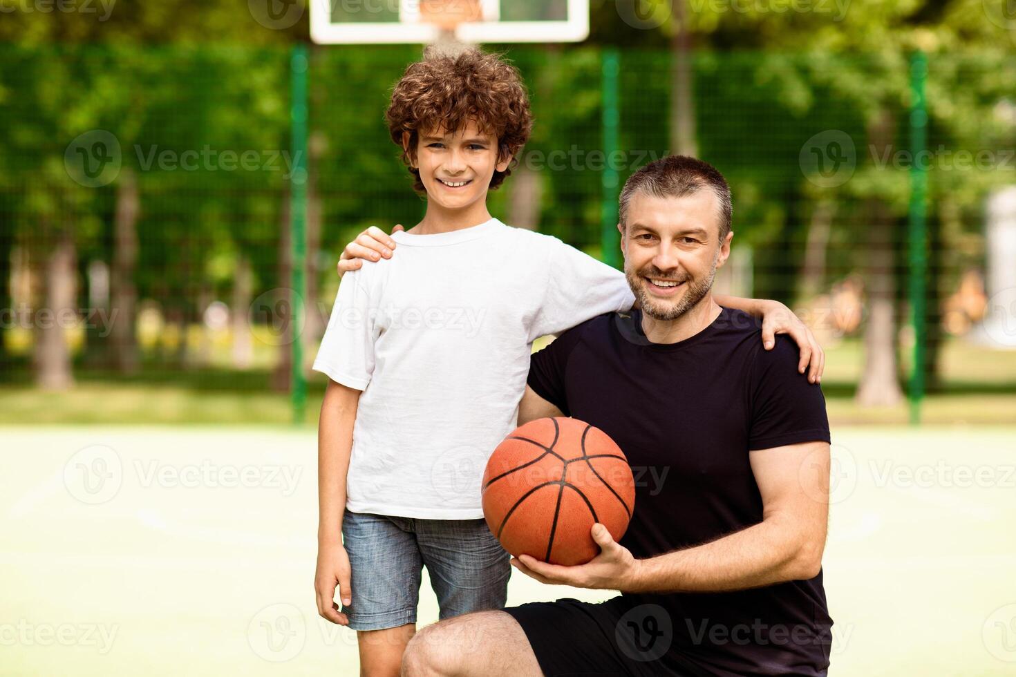 Active Lifestyle Concept. Happy boy posing with his coach on basketball field, man holding ball and looking at camera photo
