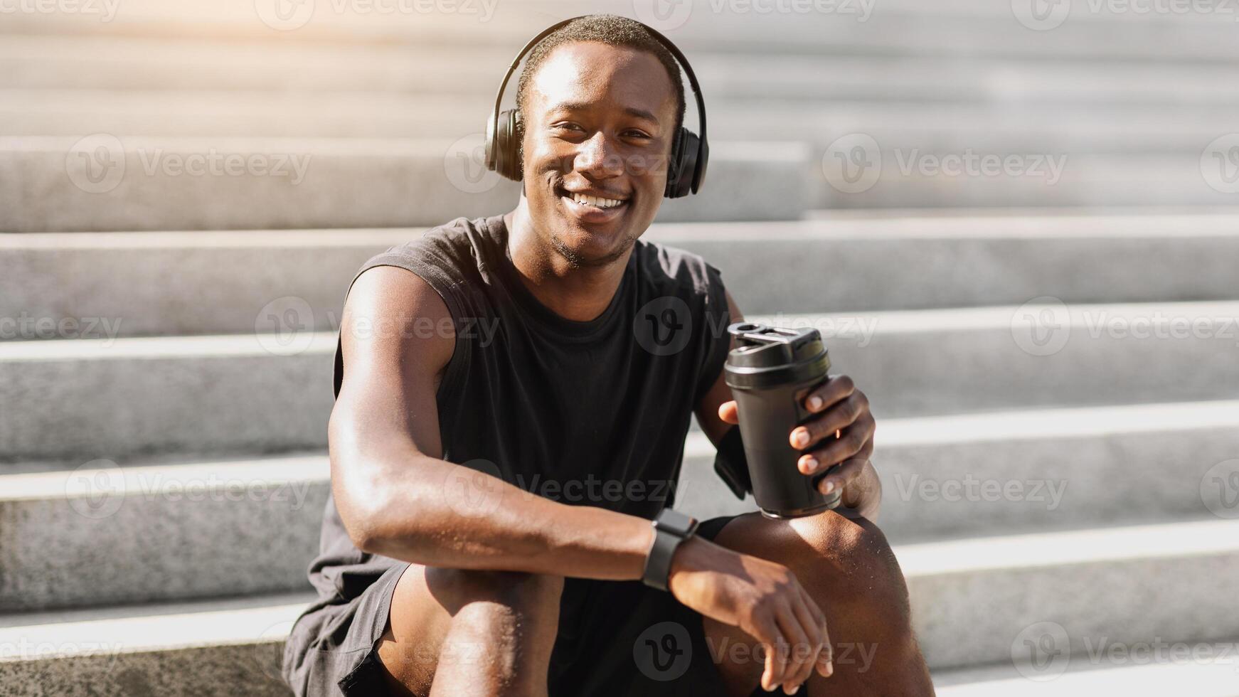 Positive Black Sportsman In Wireless Headphones Resting After Jogging Outdoors, Drinking Water, Sitting On Urban Steps And Smiling At Camera photo