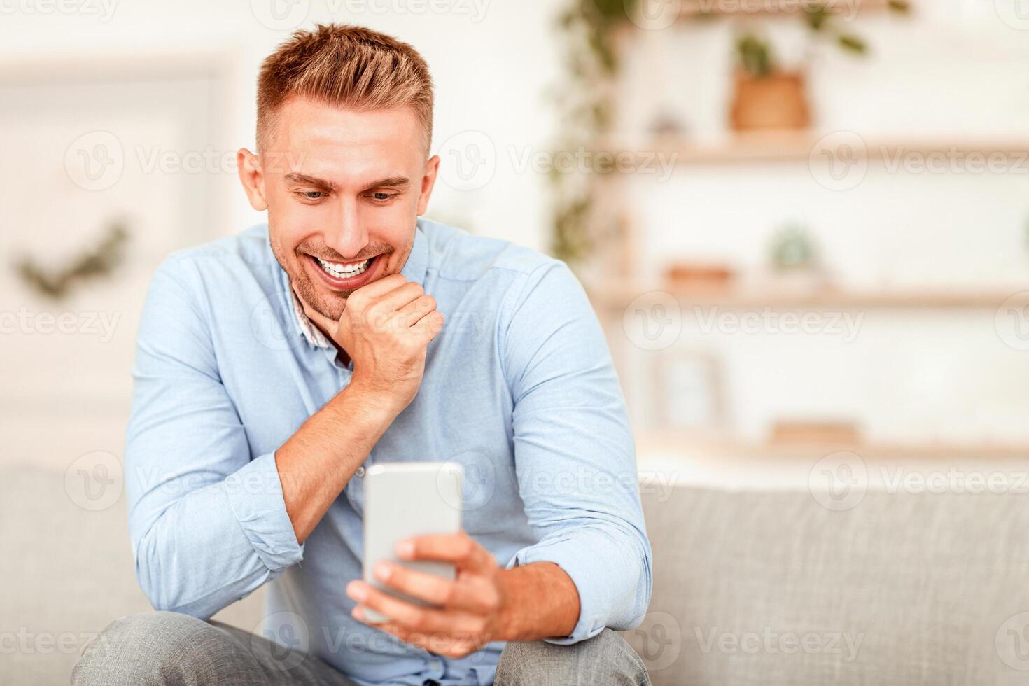 Great News. Portrait of smiling happy guy holding mobile phone, reading sms message, sitting on the sofa. Adult male browsing internet, watching , enjoying chatting and communication online photo
