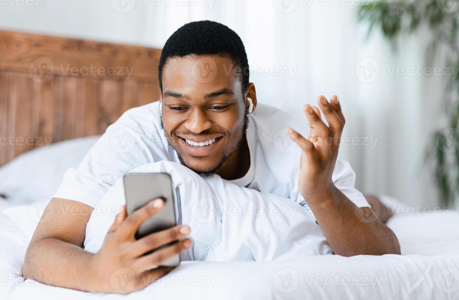Happy Black Man Making Call On Cellphone Using Smartphone And Earbuds Earphones For Distant Communication Lying In Bed In Bedroom At Home On Weekend. Selective Focus photo