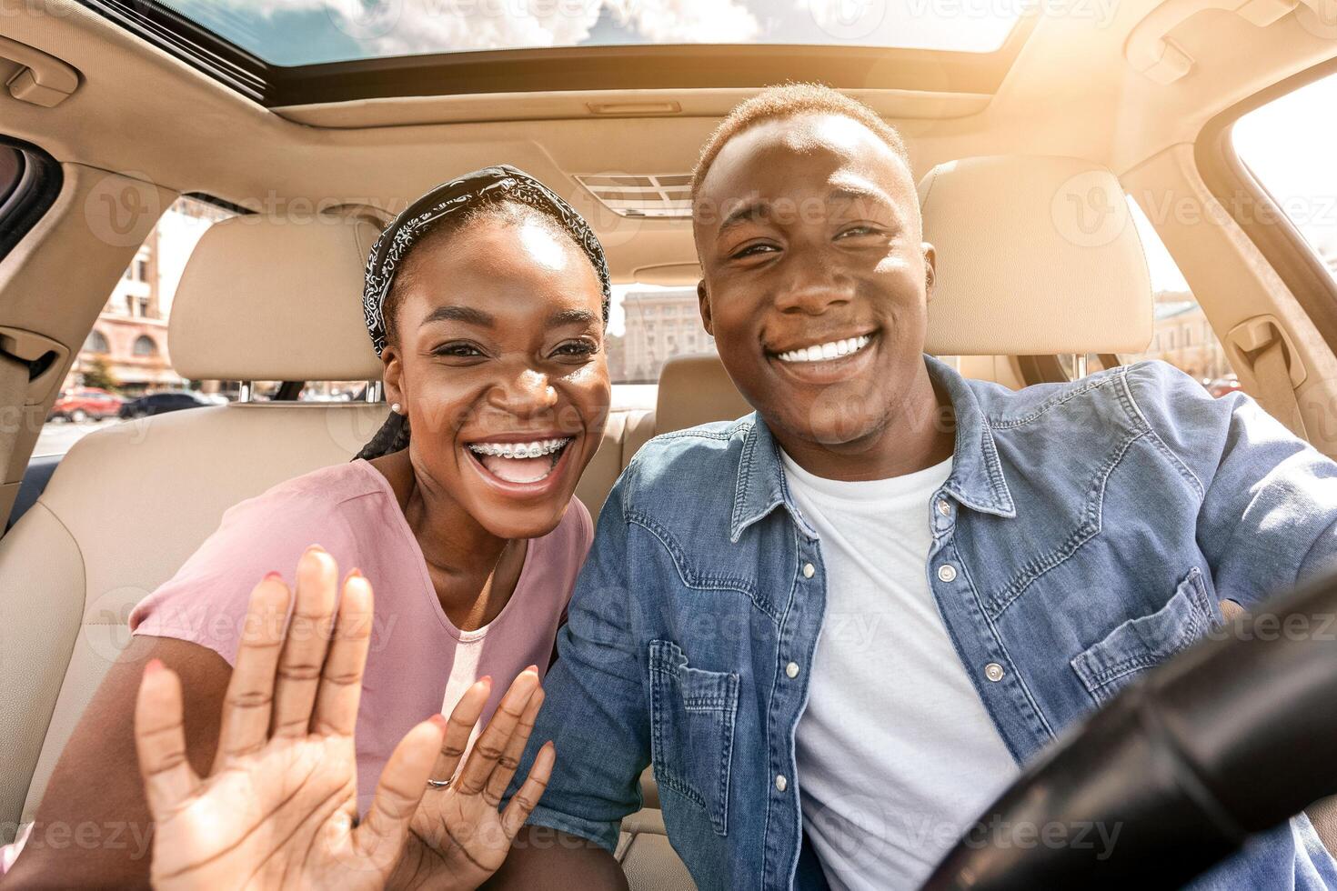 Joyful african american couple taking selfie while having car trip, sitting in auto, hugging and cheerfully smiling at camera, pretty millennial black lady embracing boyfriend and waving, sun flare photo