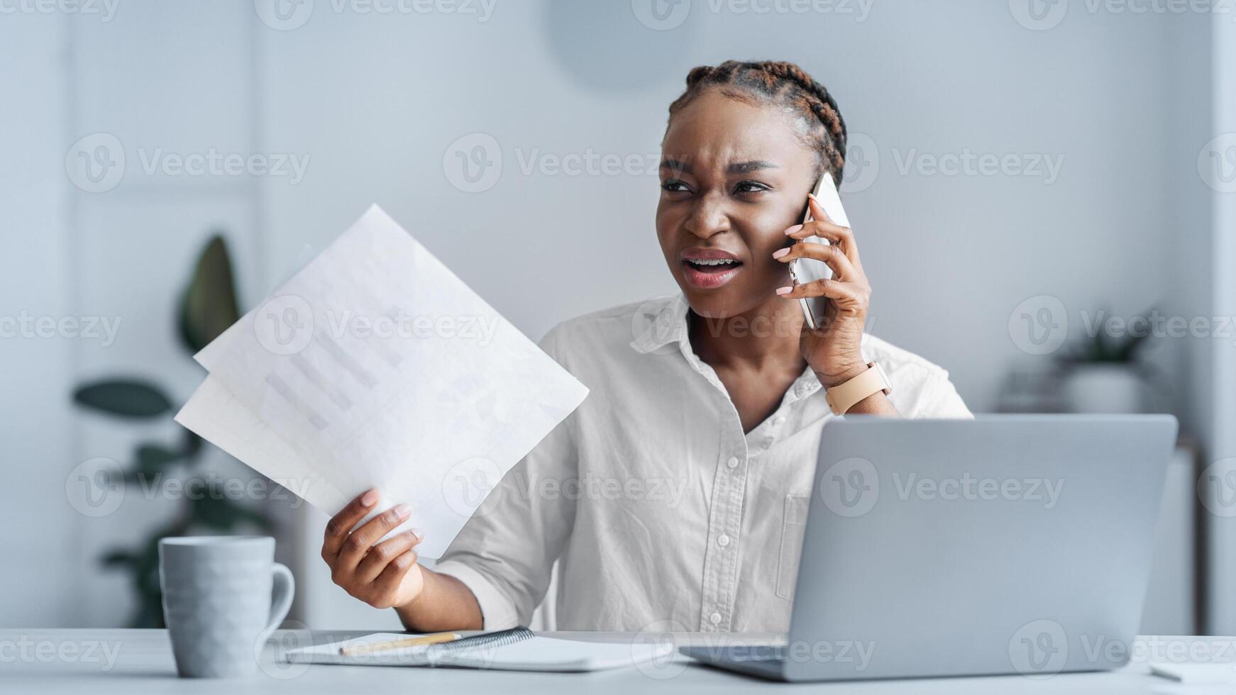 Business Stress. Angry african american businesswoman having problems in office, talking on cellphone and holding papers, arguing with client, sitting at desk with laptop at work, free space photo