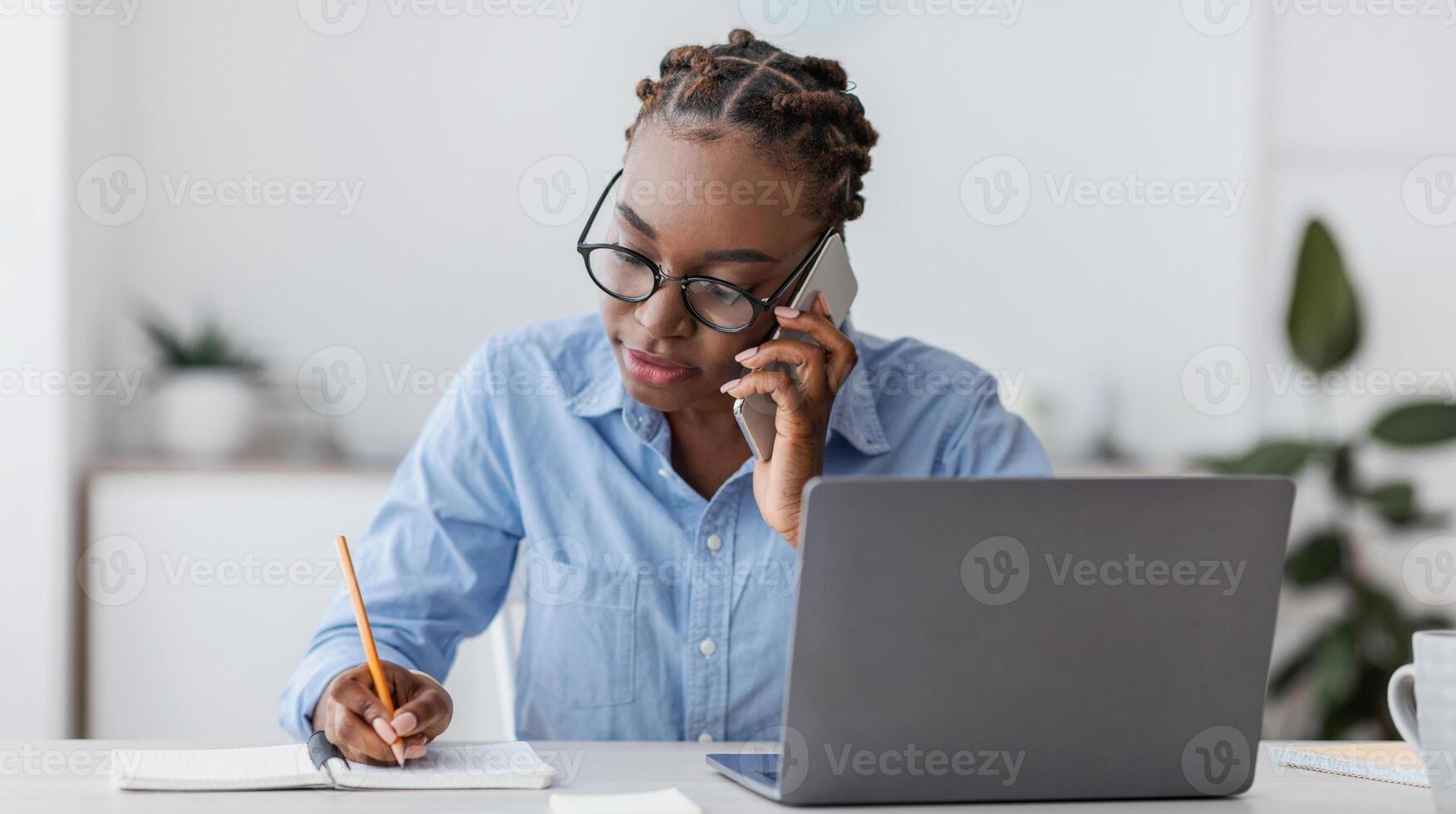 Busy Black Female Office Worker Talking On Cellphone With Client And Taking Notes, Sitting At Workplace In Modern Office, Working On Desk With Laptop, Writing Down Information To Notepad, Copy Space photo