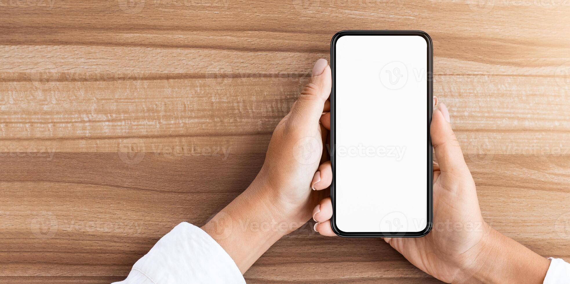 Mock up image of hand holding cell phone with isolated blank white screen, wooden table in the background photo