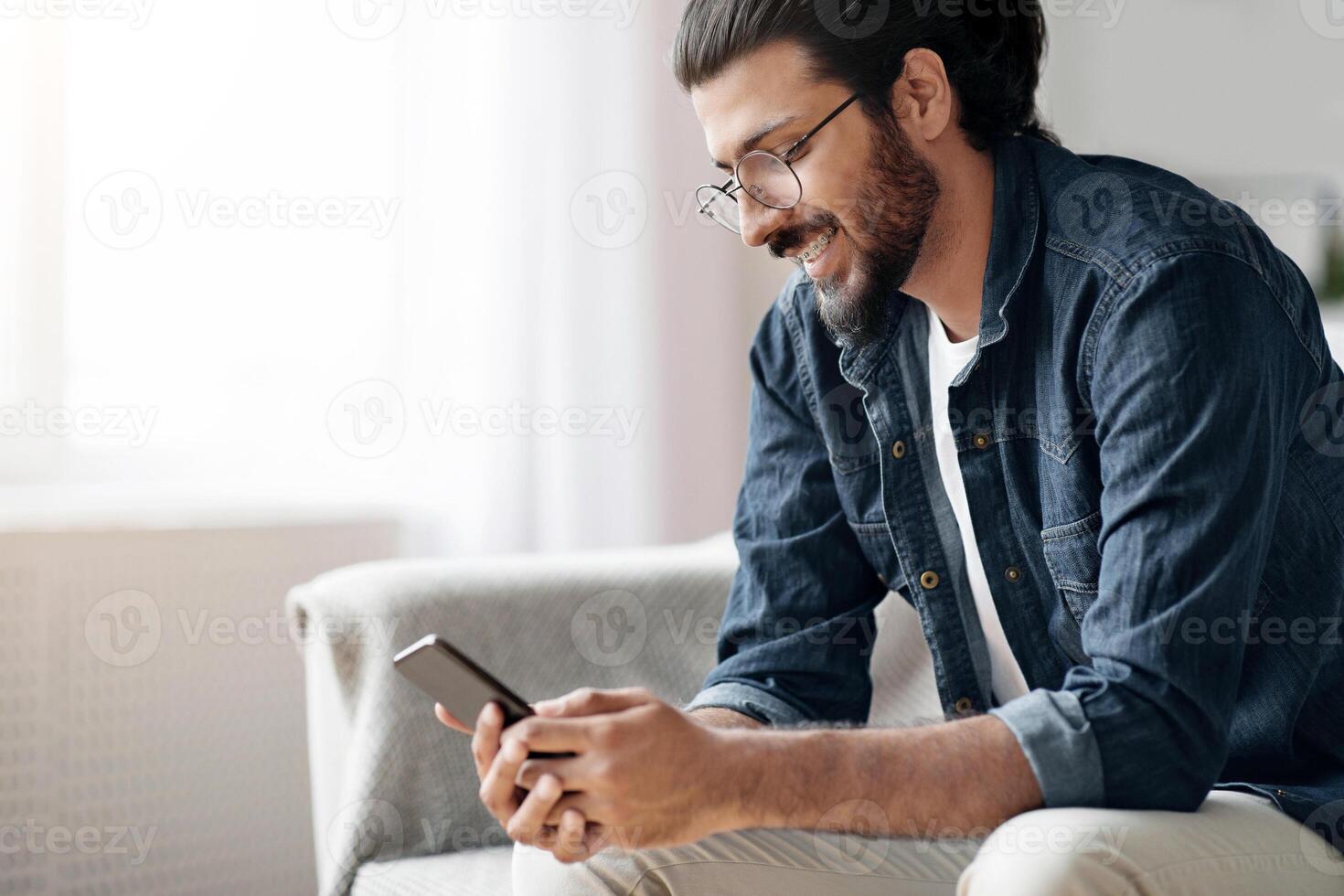 New Mobile App. Stylish Western Millennial Guy Using Smatphone While Sitting On Sofa At Home, Browsing Internet, Messaging With Friends, Playing Online Games Or Reading Sms, Wearing Glasses And Braces photo
