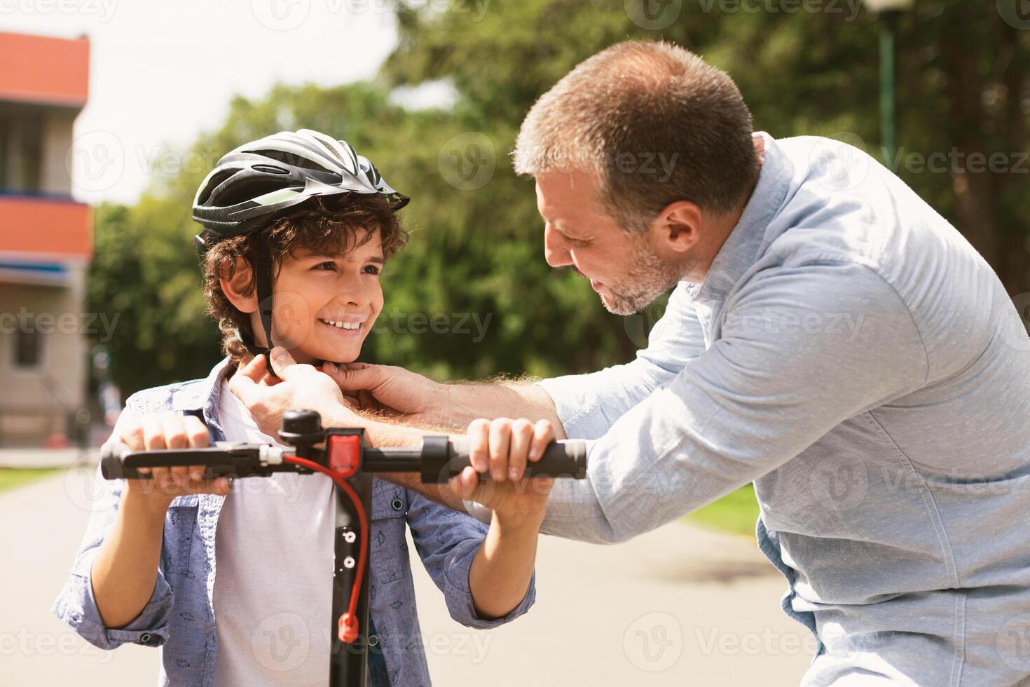 Safety Concept. Close up portrait of smiling adult father putting protection helmet on little boy at park, teaching his brave son to ride electric motorized scooter. Ourdoor Weekend Activity photo