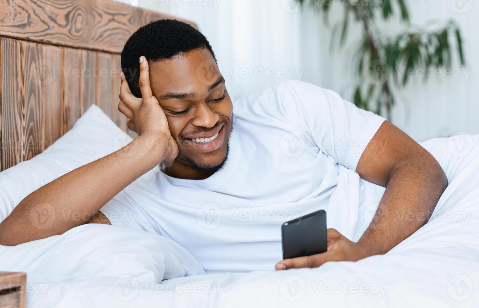 Cheerful African Man With Smartphone Browsing Internet Or Using Mobile Phone App Lying In Bed At Home. Cellphone App Advertisement, Weekend Morning Leisure Concept photo