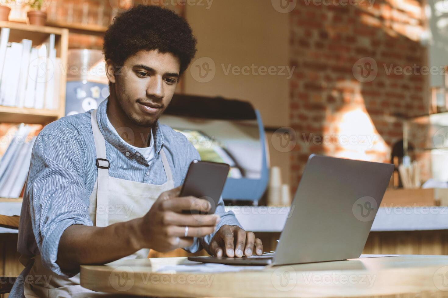 Businessman accept online orders from customers and work with social networks. Young african american man in apron looks on smartphone and works on laptop in interior of modern loft cafe, copy space photo