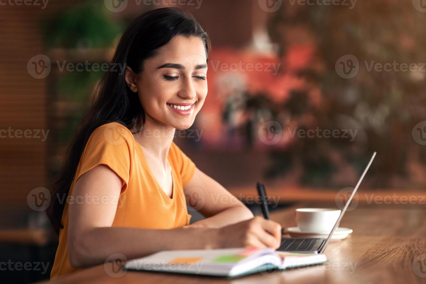 Young arab woman taking notes and websurfing at cafe, looking for job online, using laptop, copy space photo