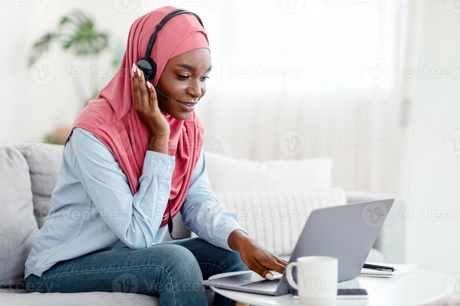 Smiling Black Muslim Female In Hijab Studying With Laptop Computer At Home, African Islamic Lady In Headscarf Using Headset, Learning Foreign Languages Online, Sitting On Couch, Copy Space photo