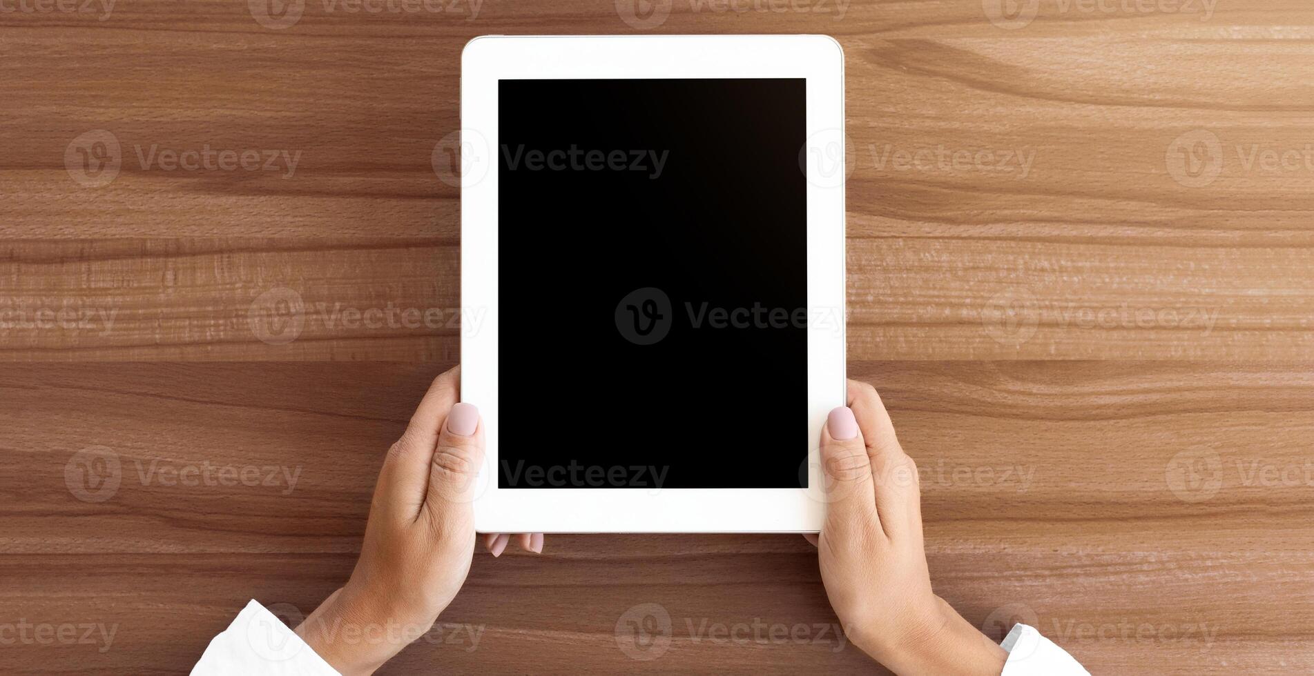 Mock up image of two hands holding digital tablet with isolated black screen, wooden table in the background photo