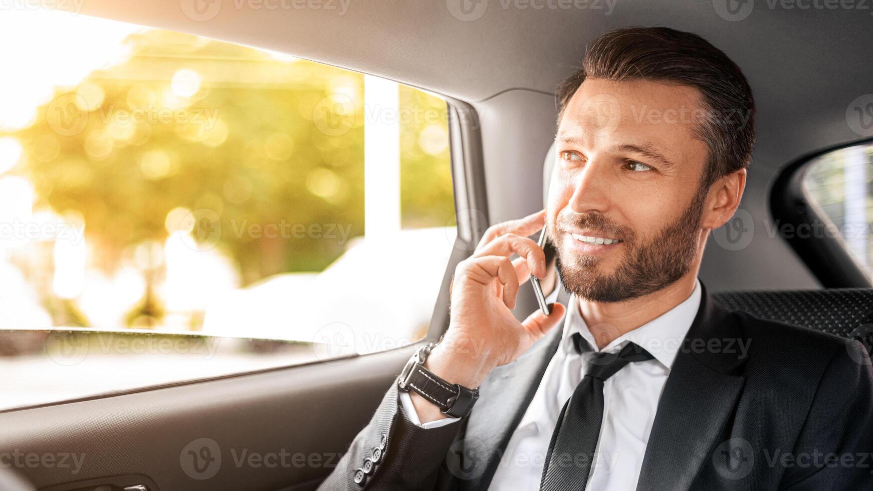Cheerful CEO or manager talking on mobile phone while going to airport by car, looking through open window. Happy businessman having business conversation while sitting in car, copy space photo