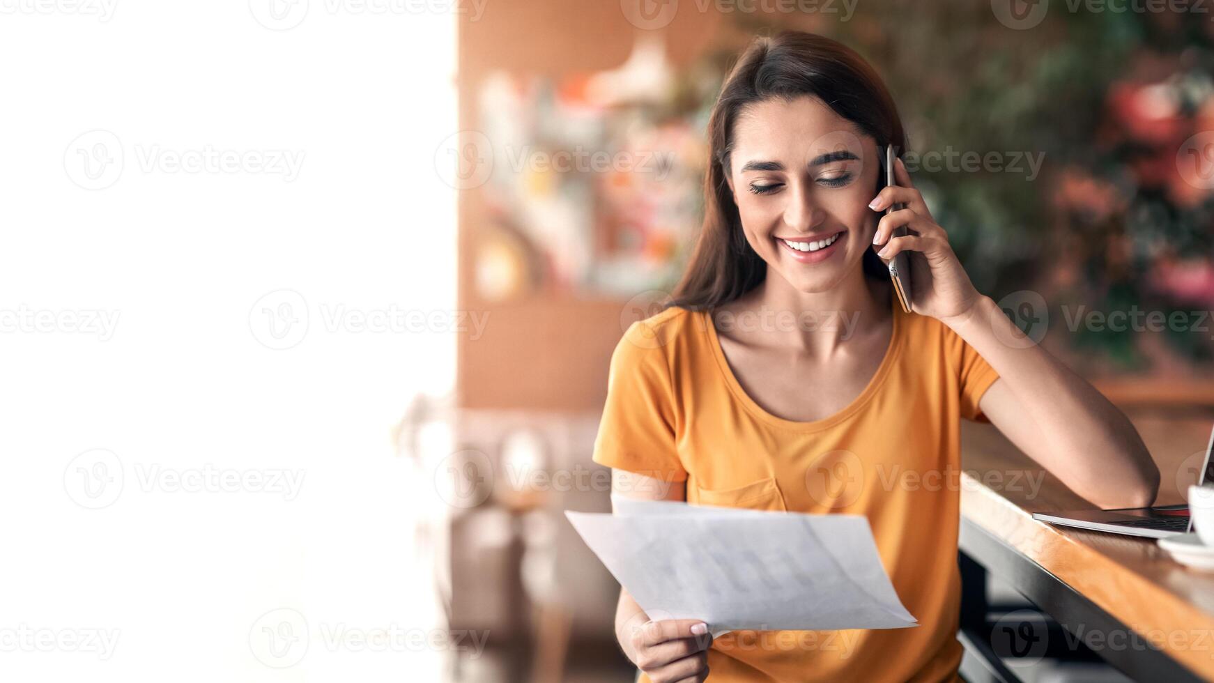 Happy arab lady having conversation on mobile phone at cafe, holding documents, working online, copy space. Smiling woman talking on smartphone with clients while drinking coffee, looking at papers photo