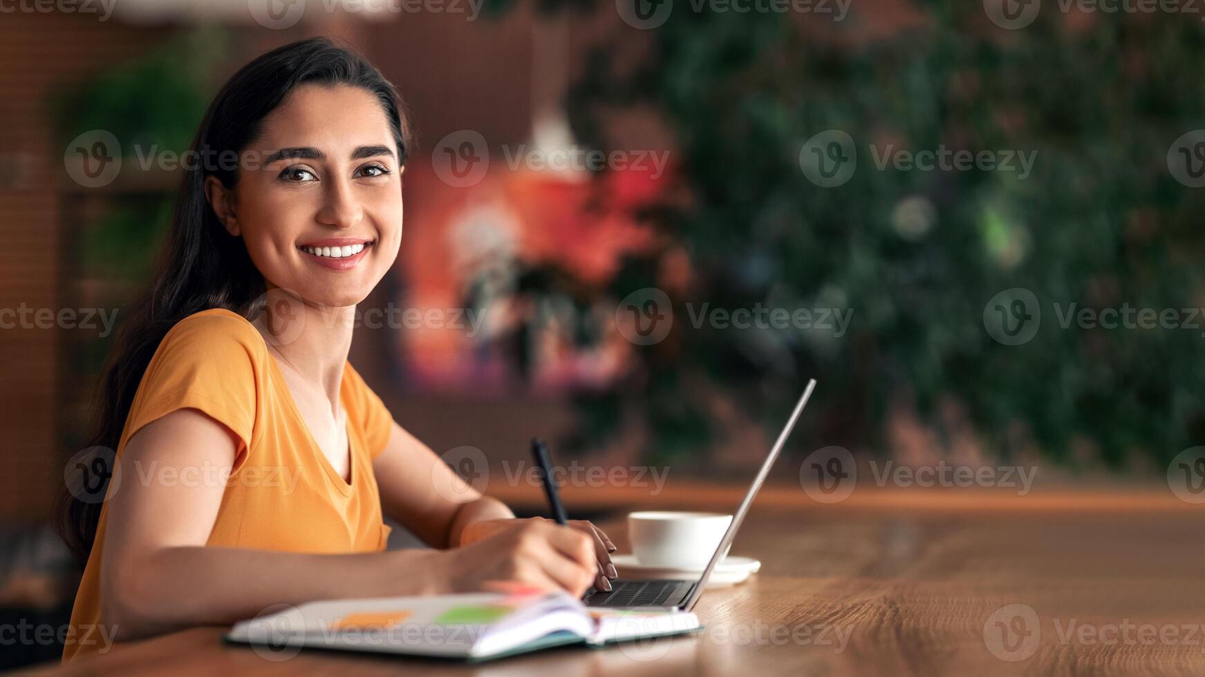 Female student looking for part-time job, taking notes and using laptop at cafeteria, free space. Young arab woman seeking job opportunities online while resting alone at cafe, side view photo