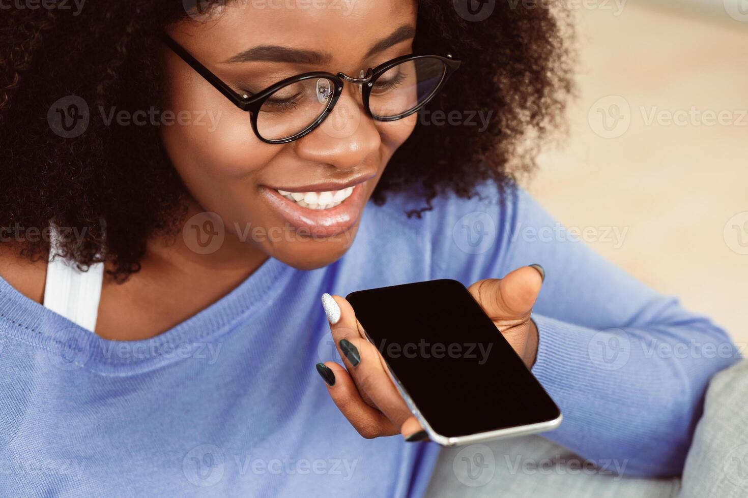 Close up of smiling black girl in glasses using smart phone voice recognition to send message, holding gadget near mouth photo